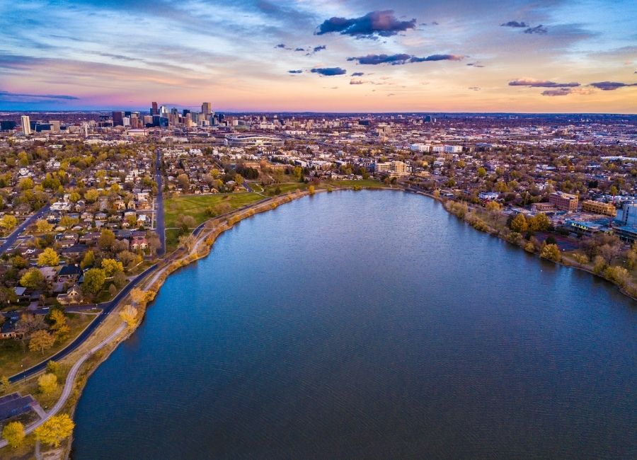 An aerial photo of Sloan Lake in Denver, Colorado, showing the large, calm lake in the foreground and the downtown Denver skyline visible in the distance under a colourful sunset sky