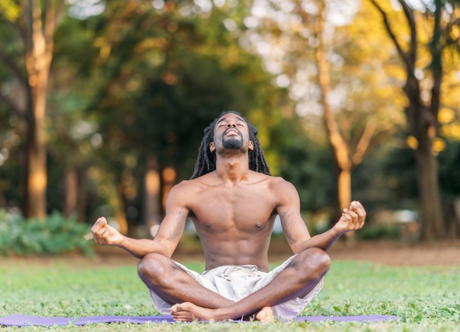Black man with long locks practising meditation outdoors on the grass surrounded by trees