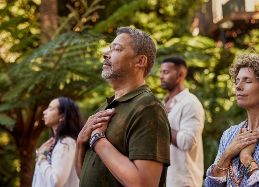 Group of adults standing together with their hands on their chests and their eyes closed in a nature setting