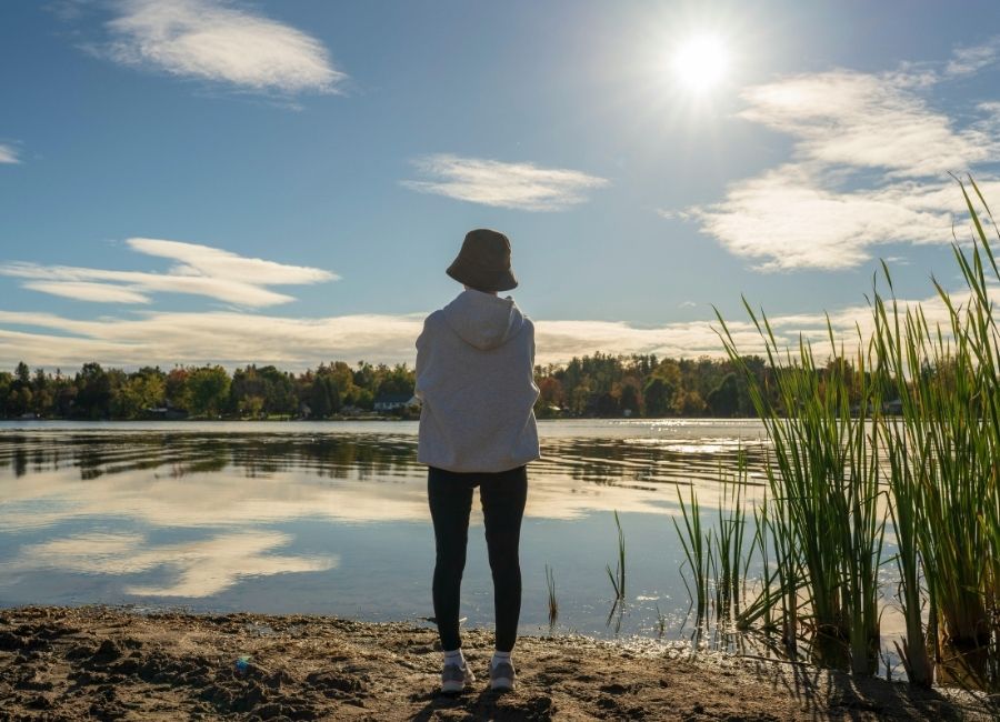 Person with a hat standing with their back to the camera in front of a lake, looking toward the water and distant trees and greenery