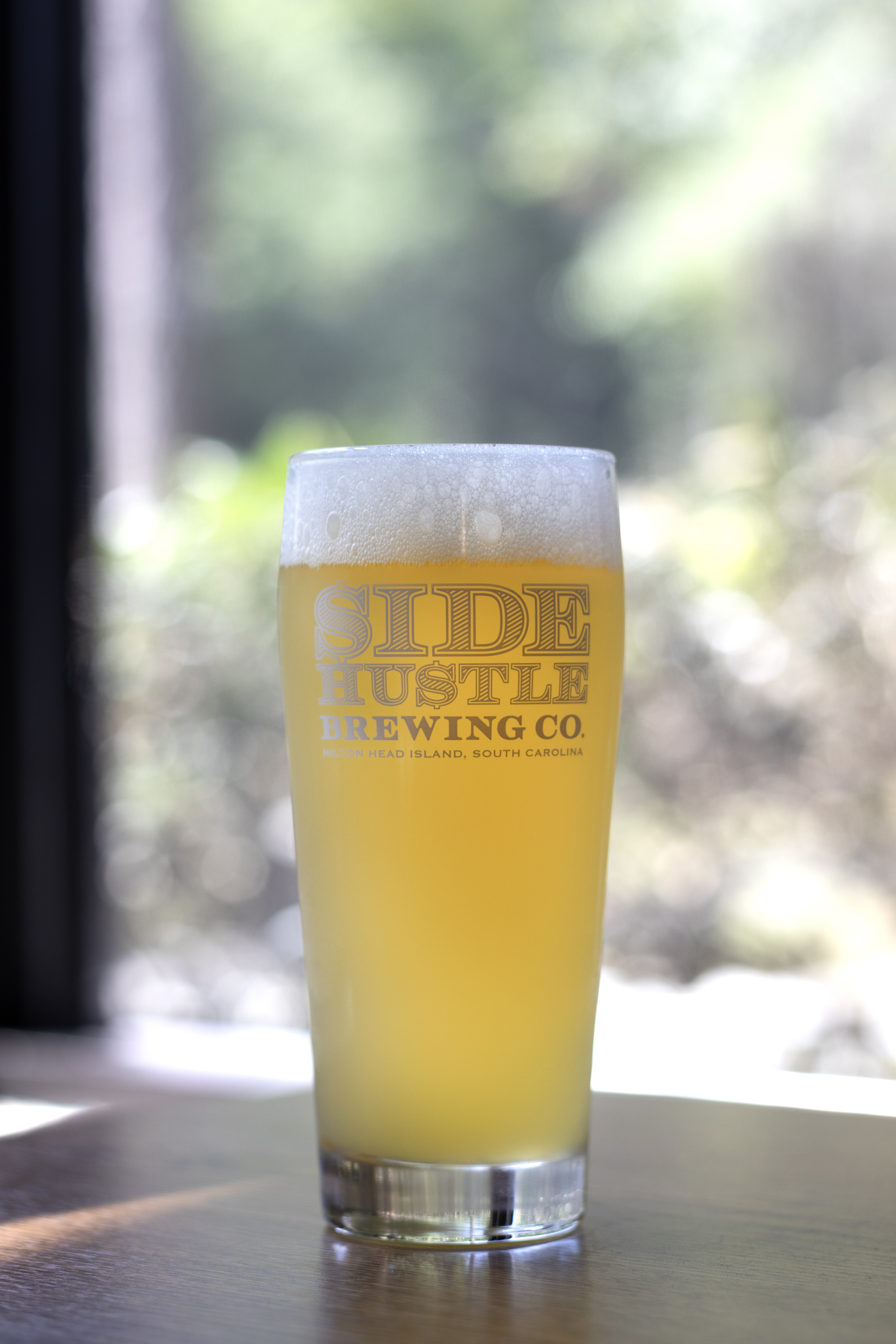 Glass of hazy pale beer with foam on top from Side Hustle Brewing Co. on a wooden table.