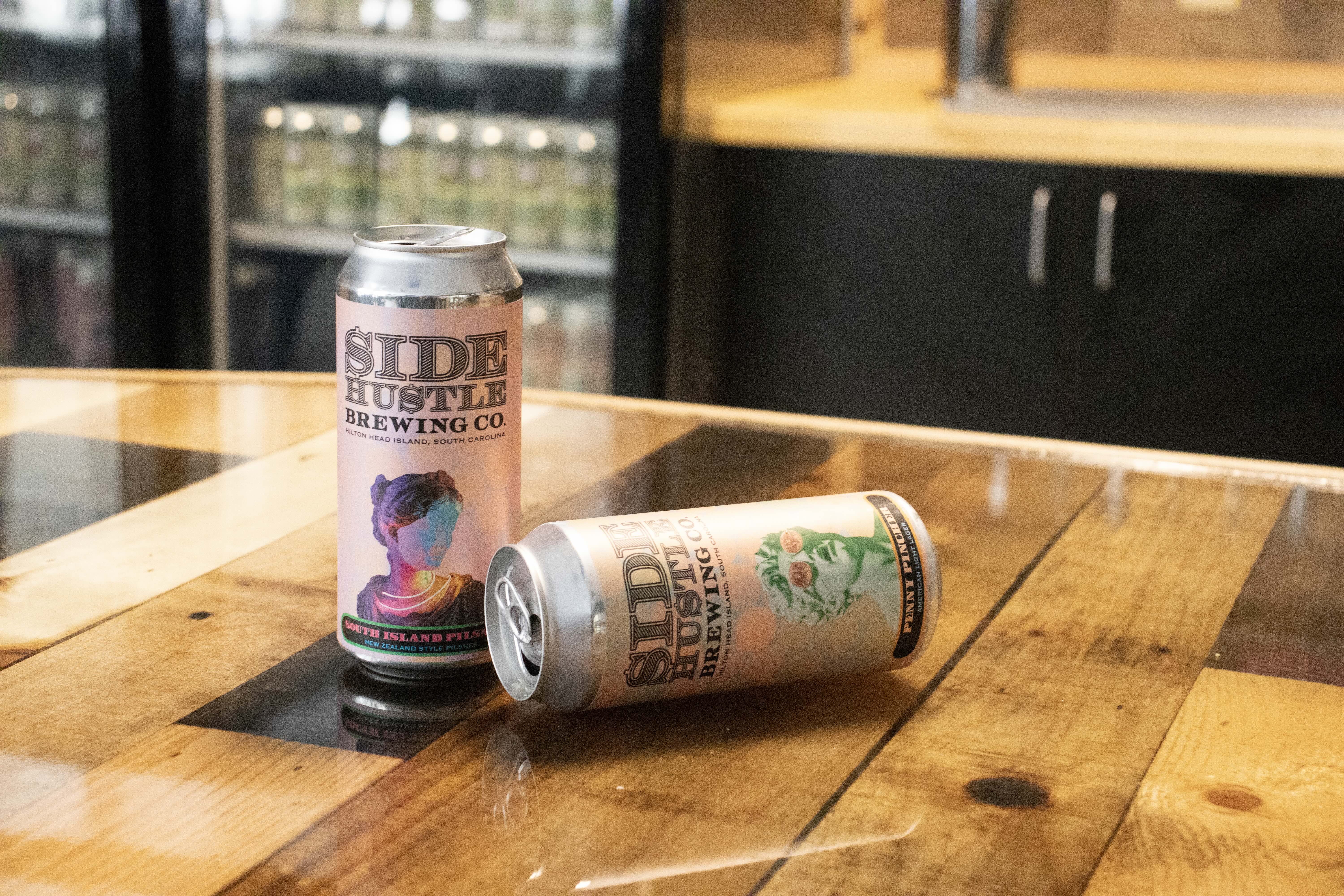 Two Side Hustle Brewing Co. beer cans on a wood table, one upright and one lying down.