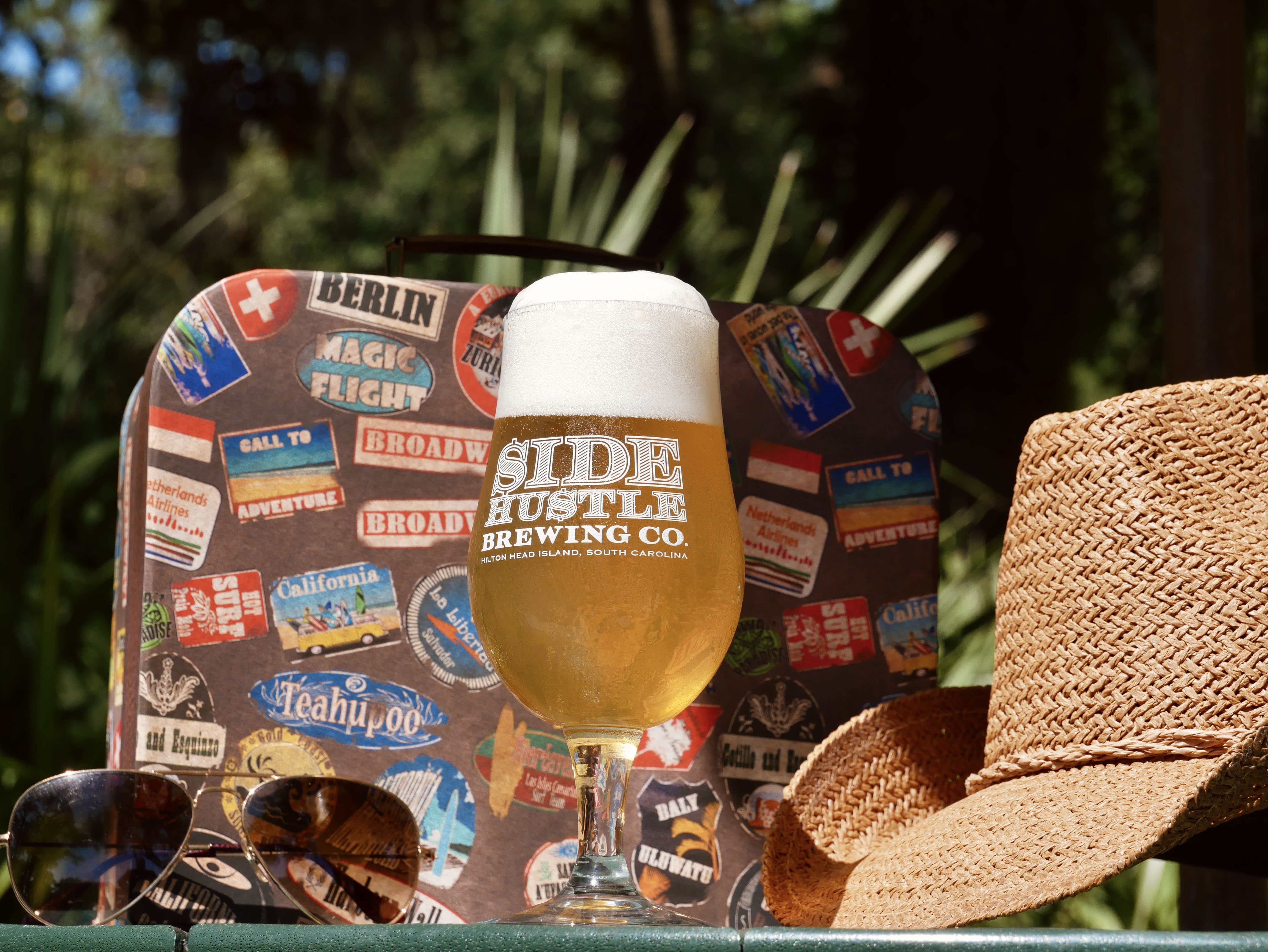 Glass of Side Hustle Brewing Co. beer with foam, sunglasses, and a woven hat outdoors.