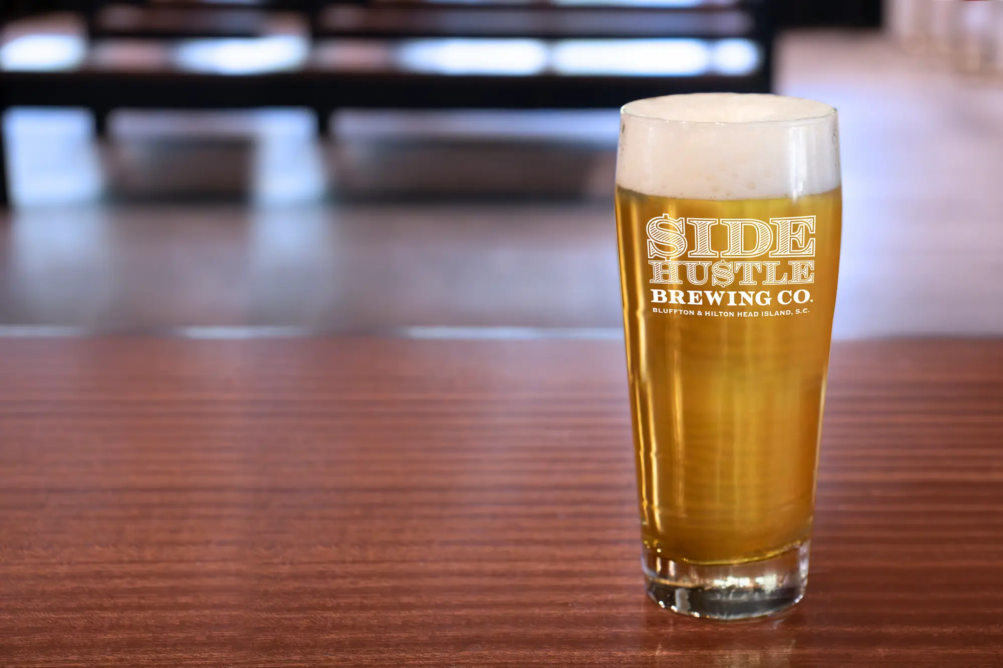 Pint glass with golden beer and foam on wooden table with Side Hustle Brewing Co. logo.