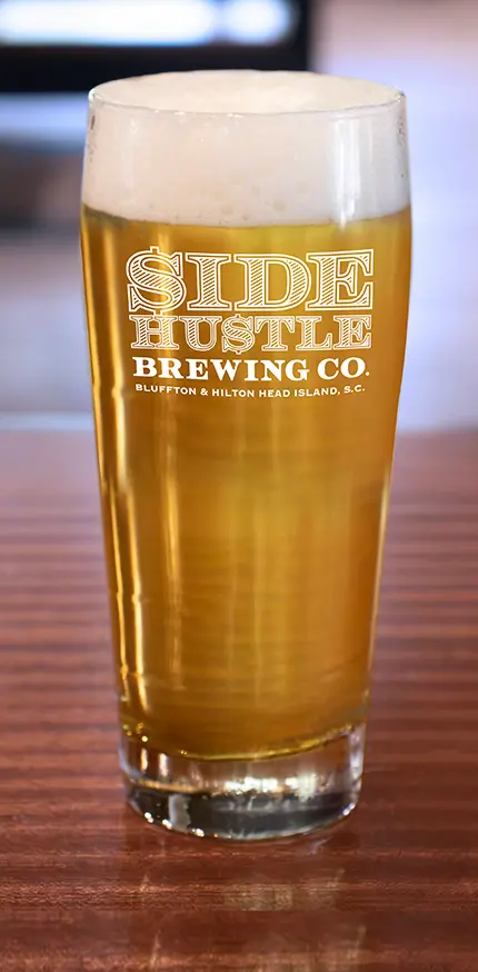 Pint glass with golden beer and foam on wooden table with Side Hustle Brewing Co. logo.
