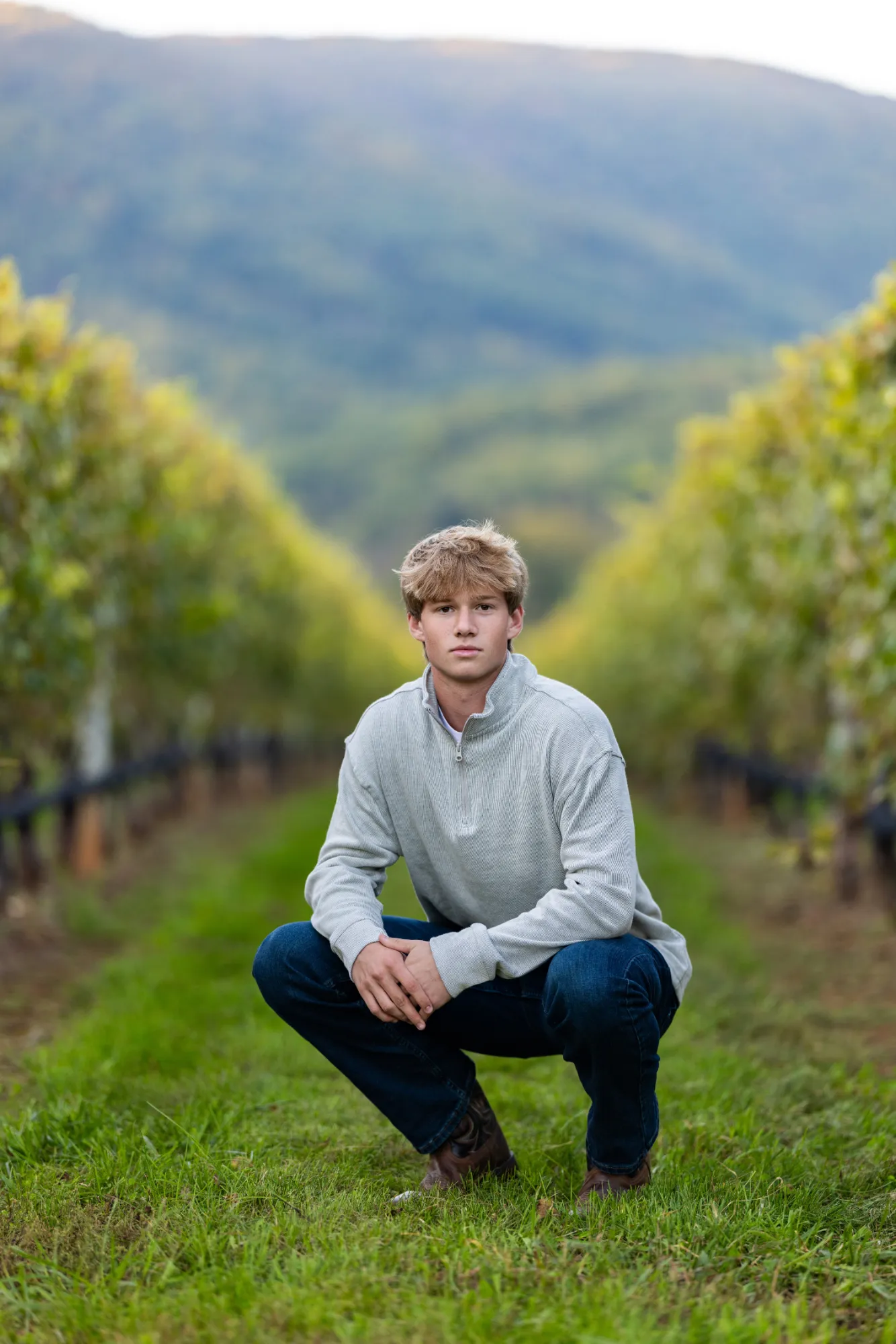 Young man in a light gray sweater and jeans kneels on grass between rows of vineyard plants with hills in the background.