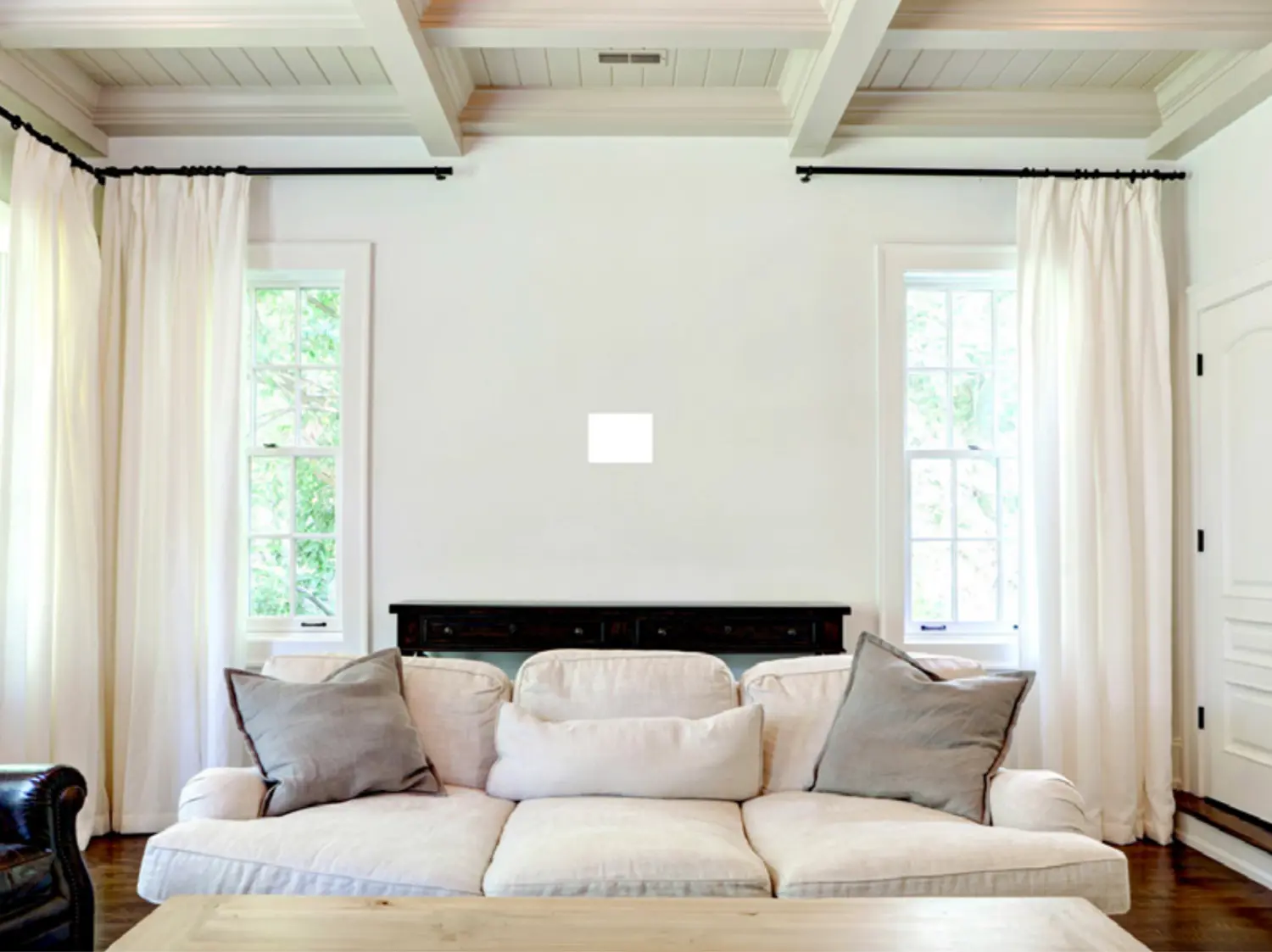 White couch with gray pillows in a bright living room with two windows and a wooden table.