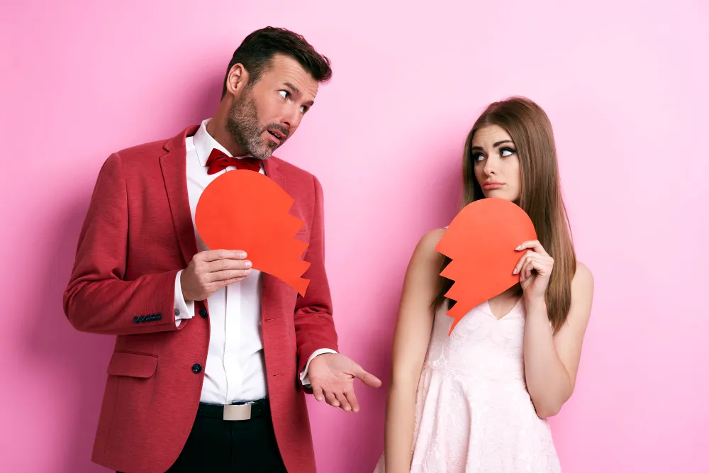 Man in red blazer and woman in white dress holding broken orange heart pieces against pink background.