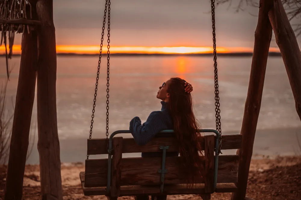 Woman sitting alone on a wooden swing at sunset by the water, reflecting on emotional readiness to date again after a breakup.