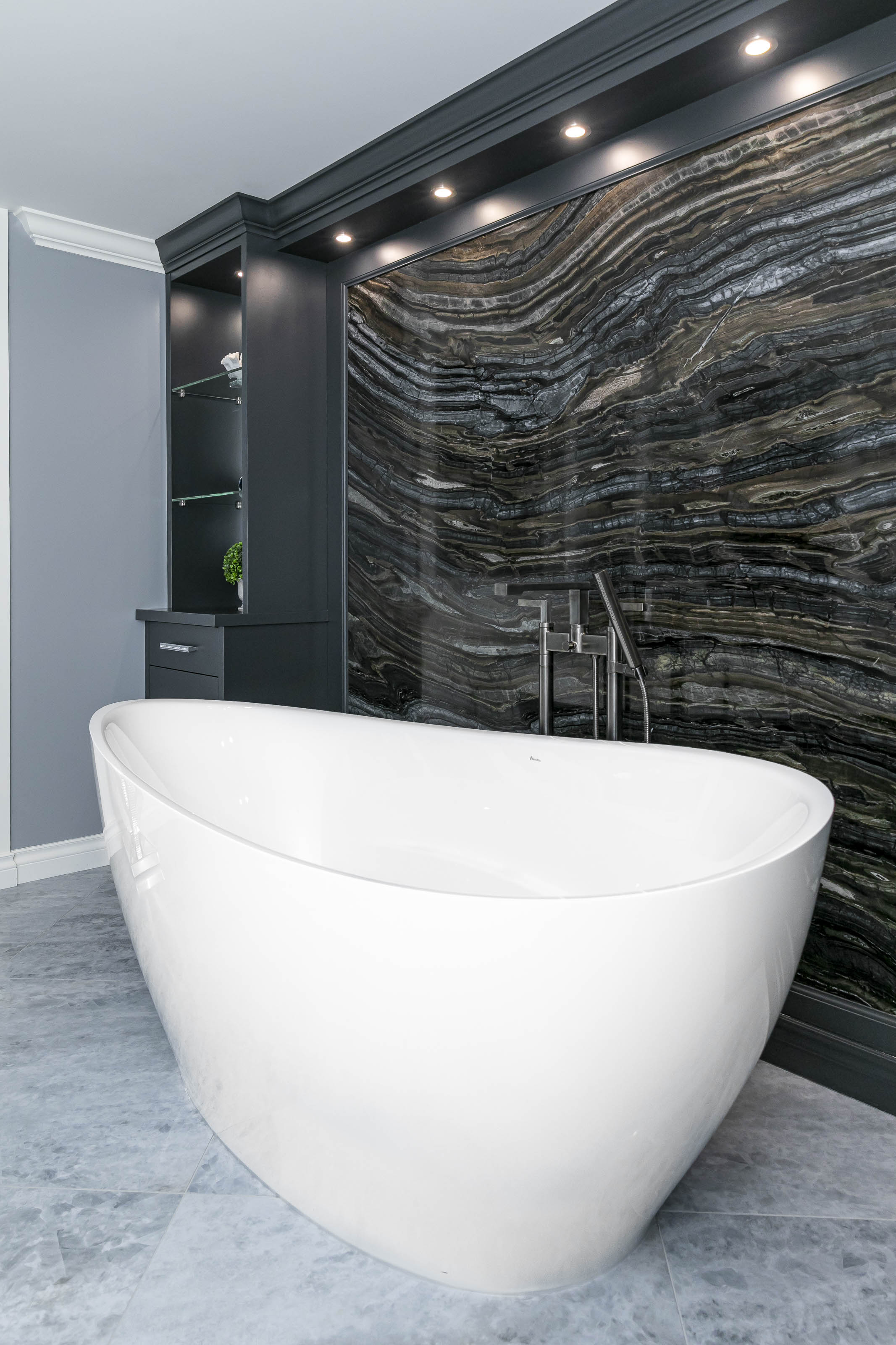 Wide shot of a freestanding white bathtub set against a striking stone slab wall and custom cabinetry, part of a residential bathroom build in Victoria, BC.