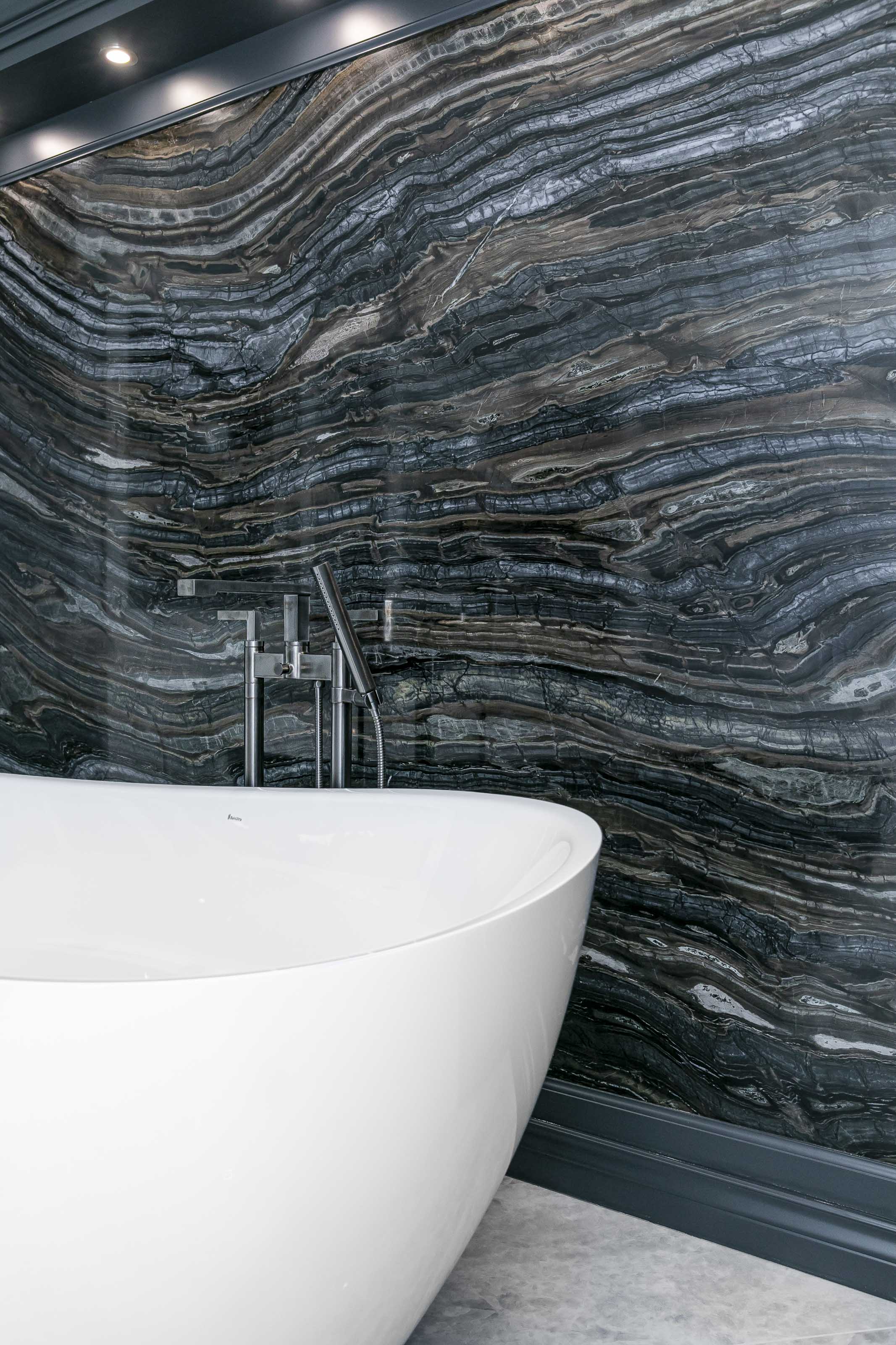 Close-up of a luxurious soaker tub with a floor-mounted faucet in front of a dramatic, veined stone feature wall in a high-end bathroom designed by Outlook Project Management.