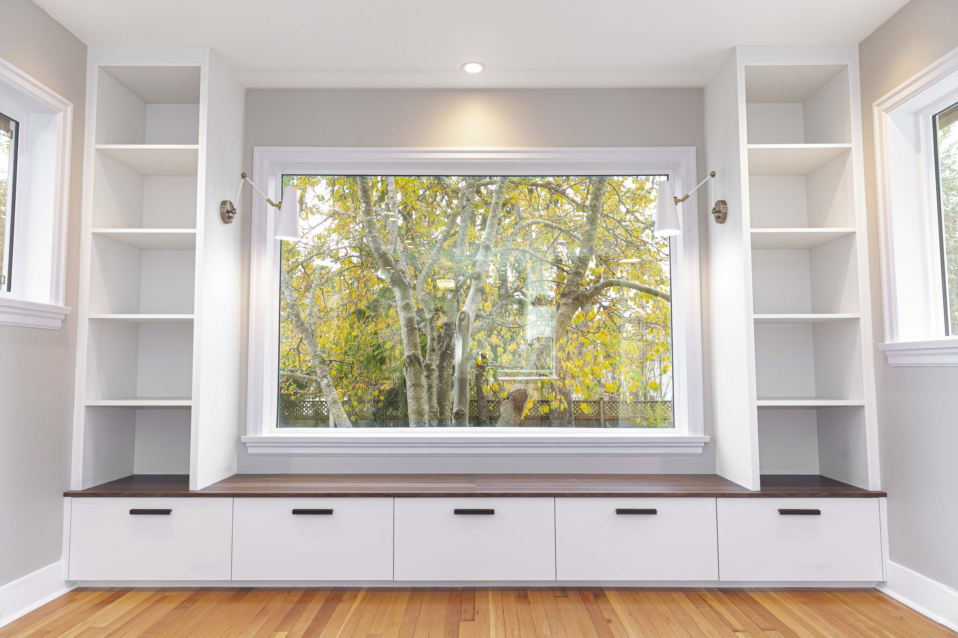Built-in white window seat with storage drawers, open shelving, and a large picture window showcasing a tree with autumn leaves.