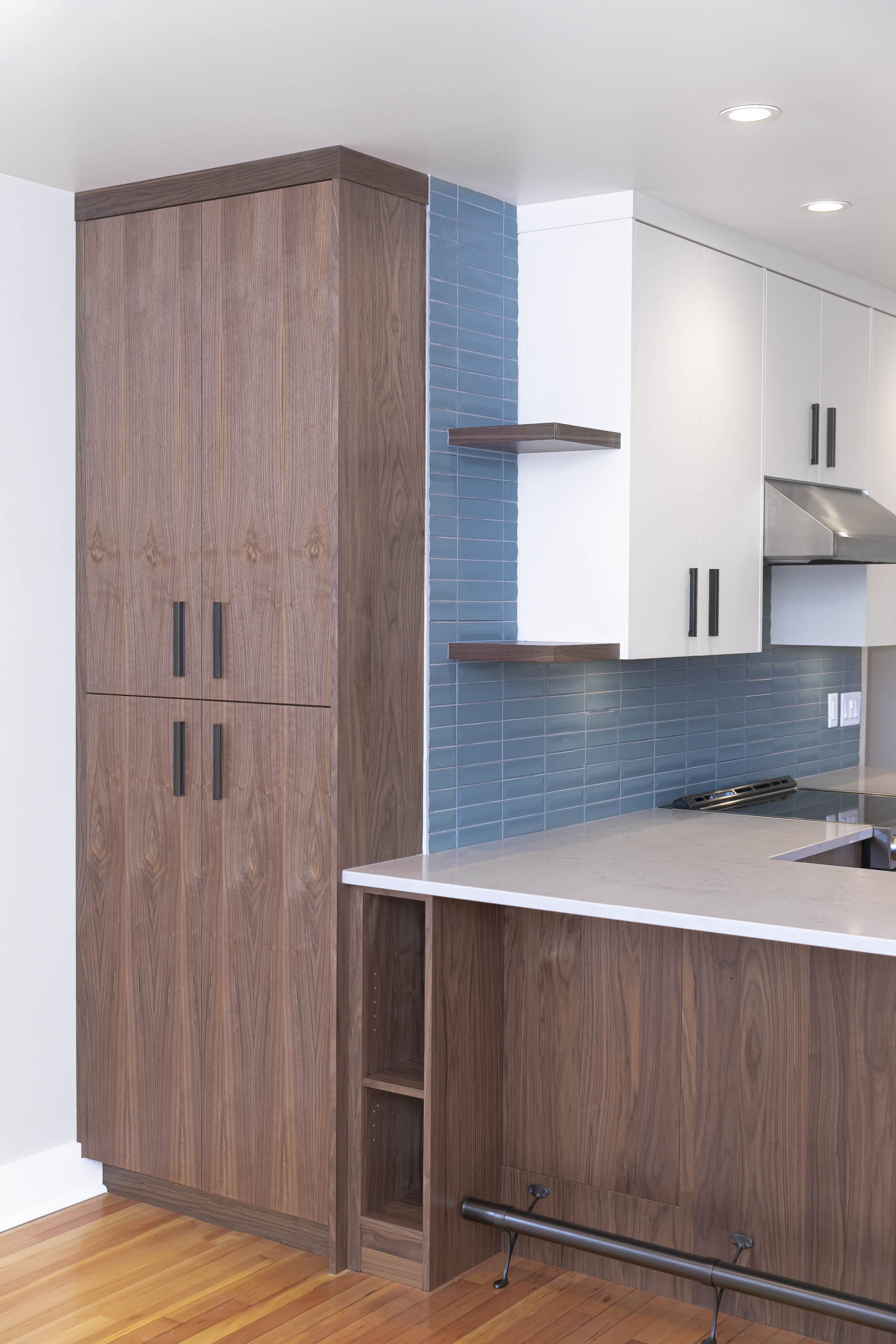 Wide view of a renovated kitchen with white upper cabinets, walnut base cabinetry, open shelving, and a full blue tile backsplash.