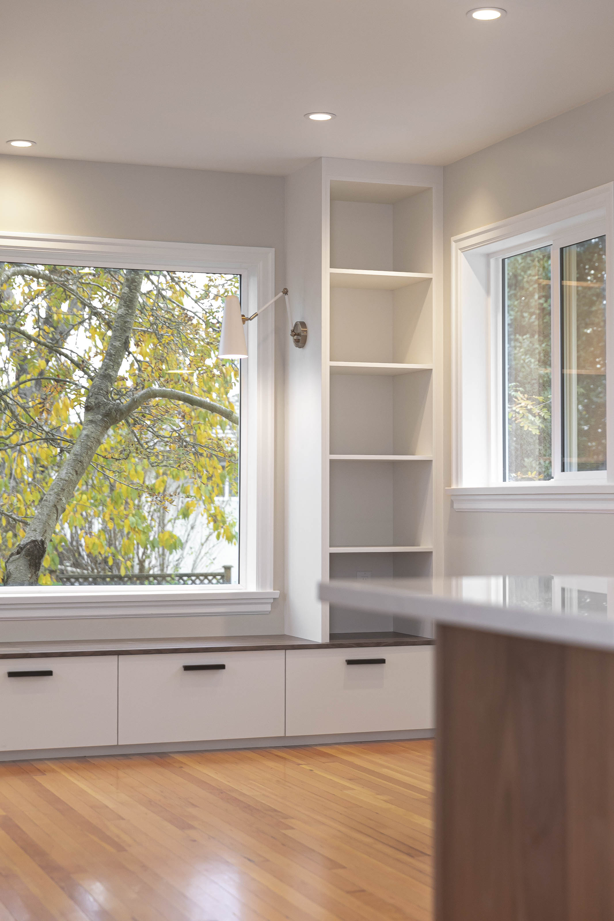 Corner angle of a cozy reading nook with open white bookshelves, bench drawers, and two large windows looking out at autumn foliage.