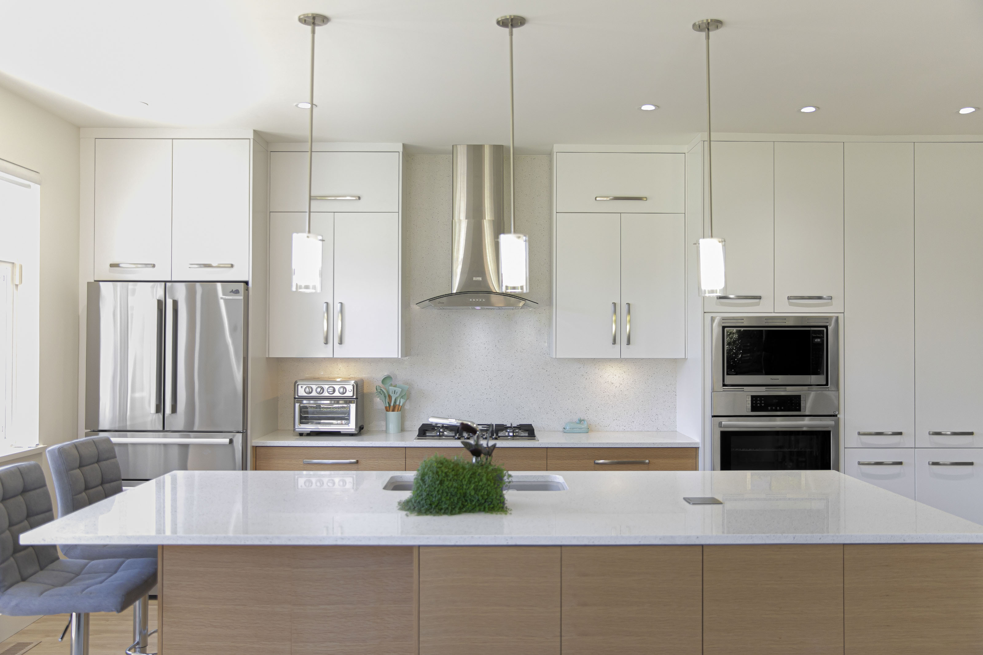 Modern kitchen renovation featuring white upper cabinets, natural wood lower cabinets, stainless steel appliances, and pendant lighting above a large quartz island.