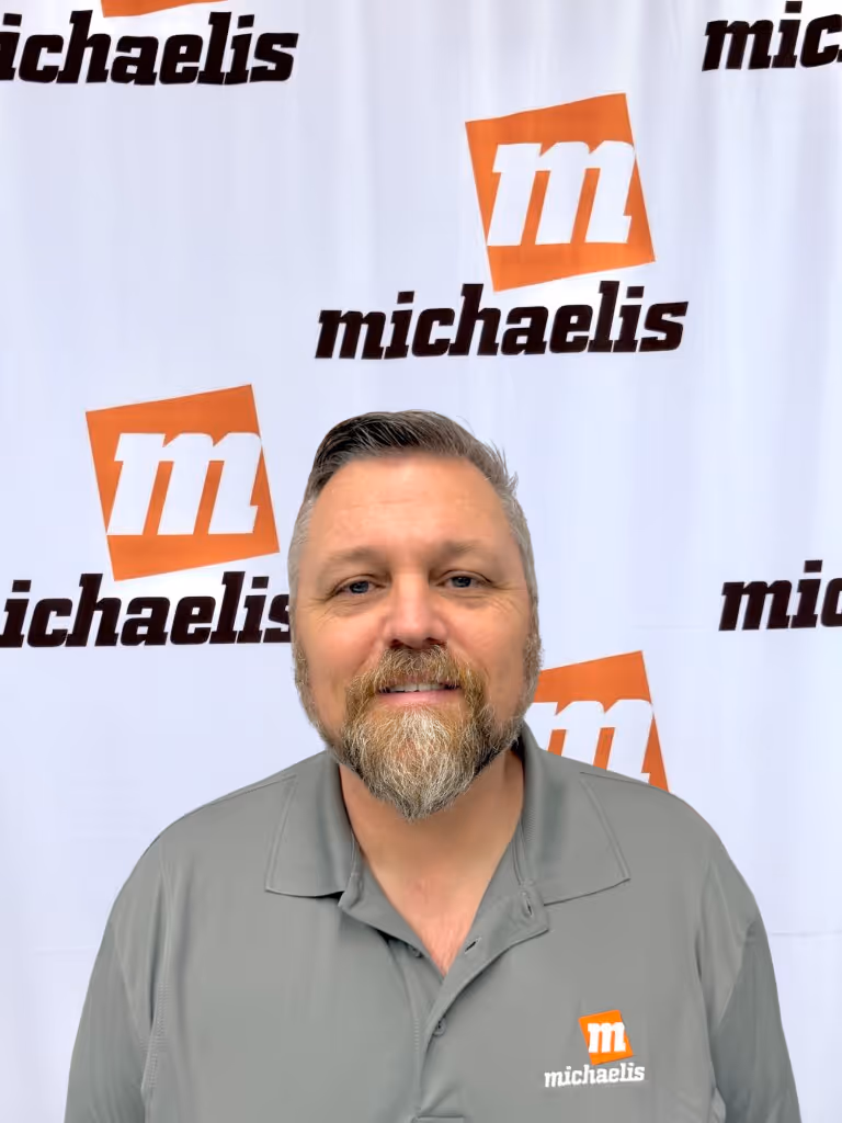 Man with a beard wearing a gray polo shirt with the Michaelis logo, standing in front of a Michaelis branded backdrop.