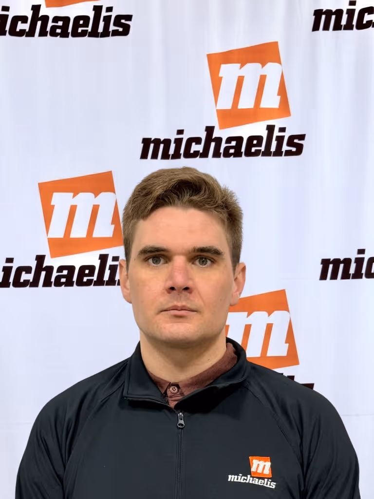 Man with short brown hair wearing a black zip-up jacket with the Michaelis logo, posing in front of a white background with repeated Michaelis logos.