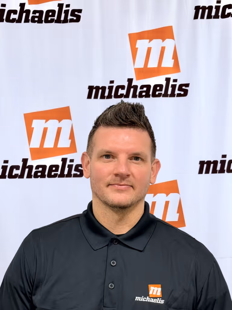 Man with short spiked hair wearing a black polo shirt with the Michaelis logo, standing in front of a Michaelis branded backdrop.