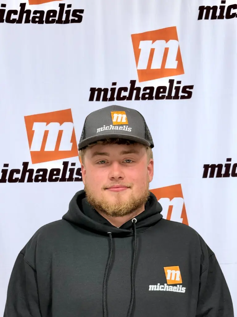Man with blonde beard wearing a black hoodie and black cap, both branded with 'michaelis' logo, standing in front of a backdrop displaying the same logo.
