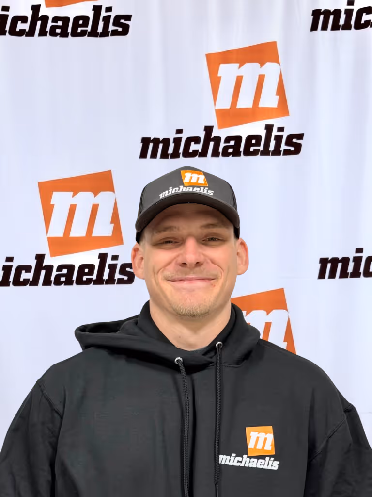 Smiling man wearing a black hoodie and cap with Michaelis logo, standing in front of a Michaelis branded backdrop.