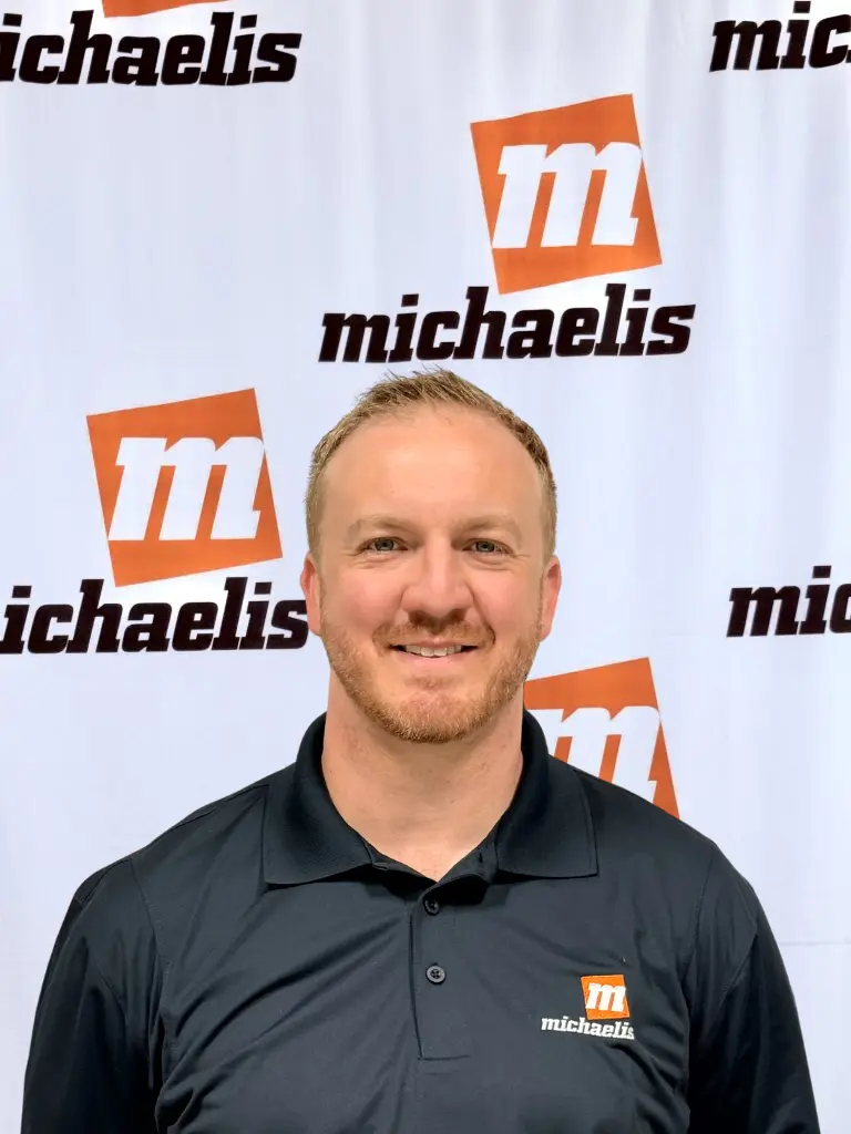 Young man with curly hair wearing a black Michaelis polo shirt in front of a Michaelis-branded backdrop.
