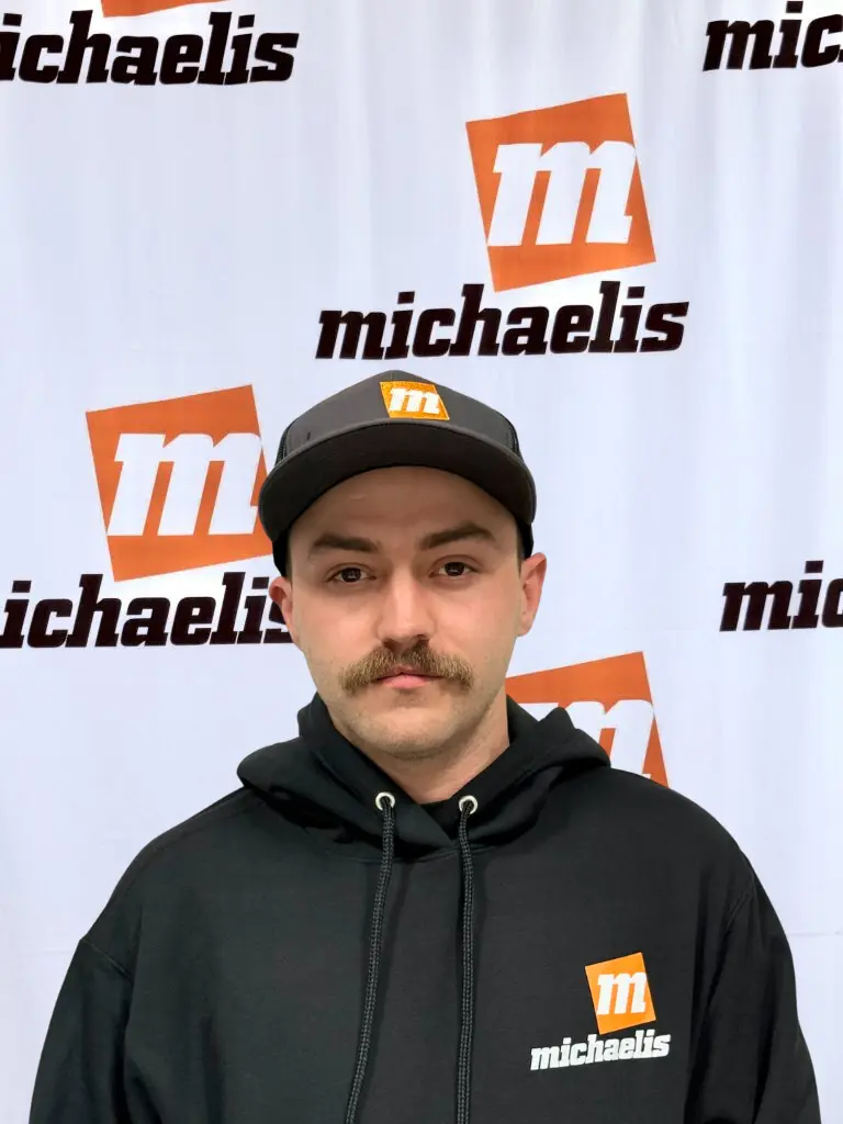 Young man wearing an orange shirt and black cap with the michaelis logo standing in front of a michaelis branded backdrop.