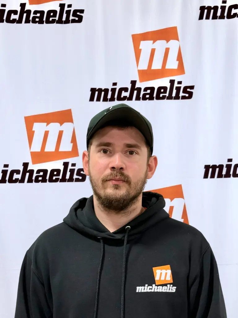 Young man with light brown hair wearing an orange Michaelis shirt standing in front of a white backdrop with Michaelis logos.