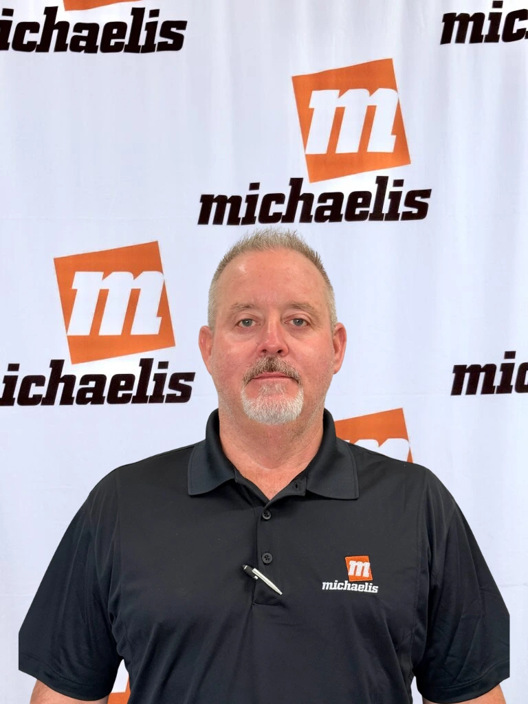 Man with a beard wearing a gray polo shirt with the Michaelis logo, standing in front of a Michaelis branded backdrop.