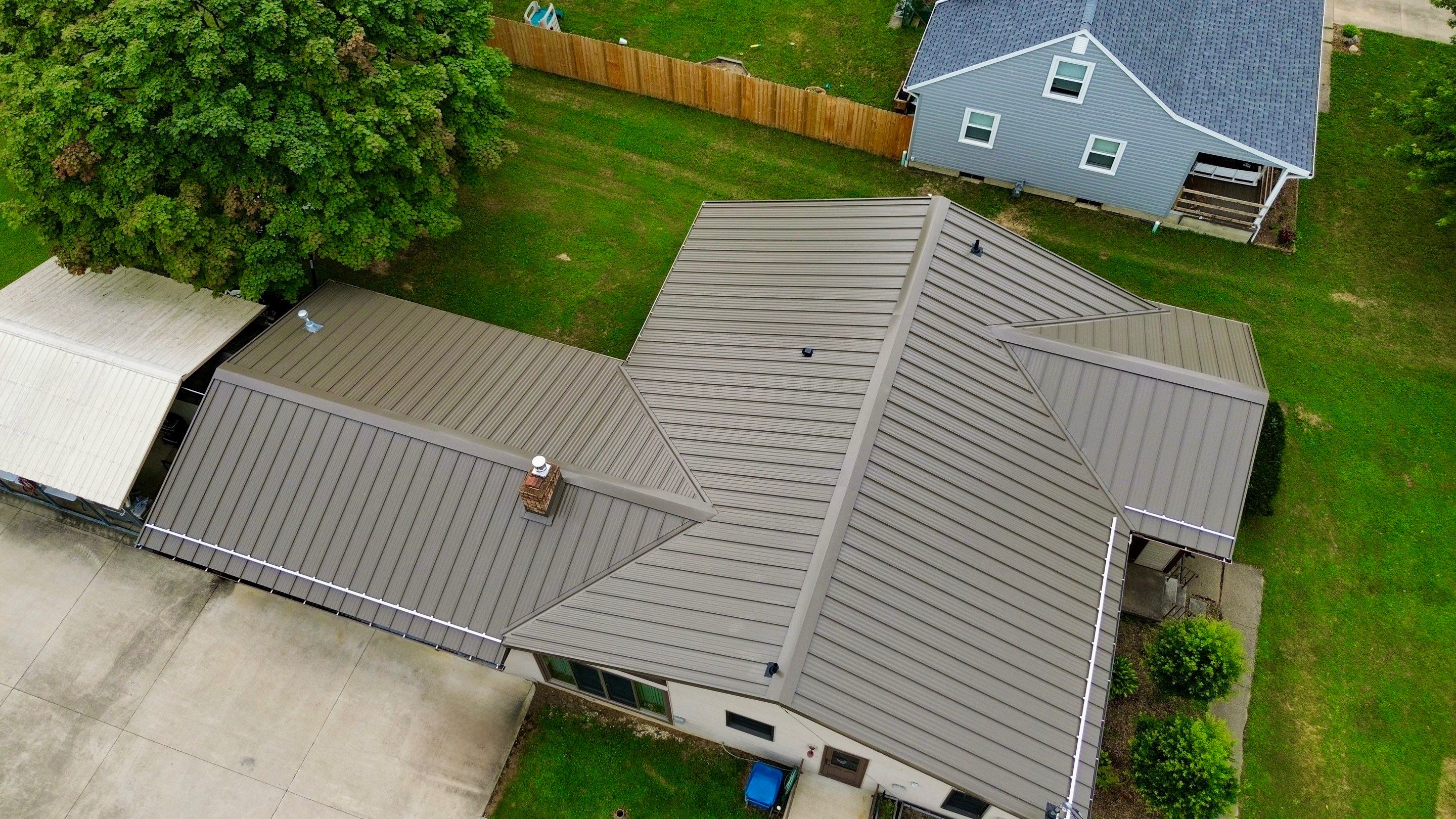 Aerial view of a house featuring a brown metal roof, surrounded by green grass and a wooden fence.