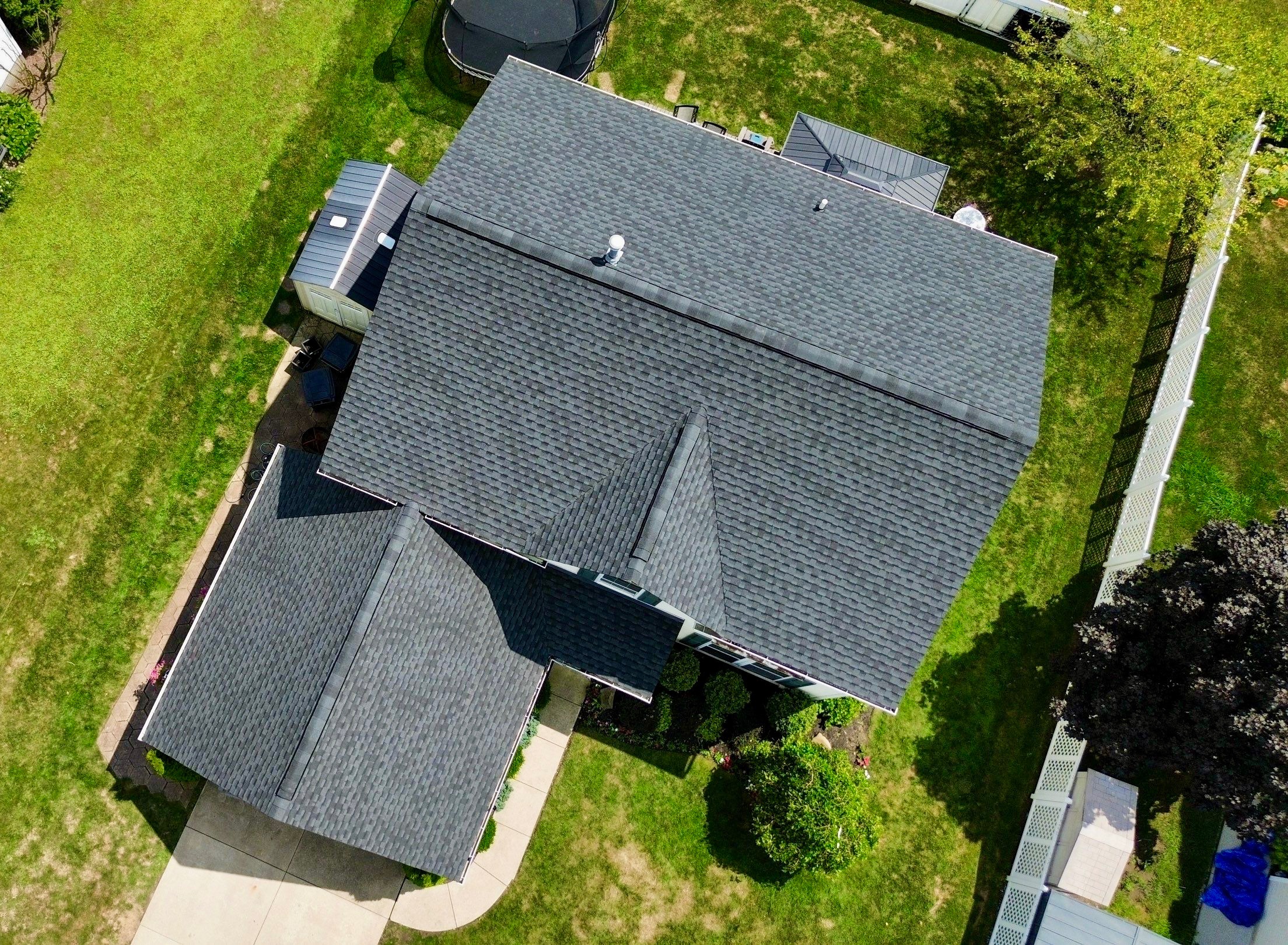 An aerial view of a house with a multi-pitch roof surrounded by green grass and a fenced yard.