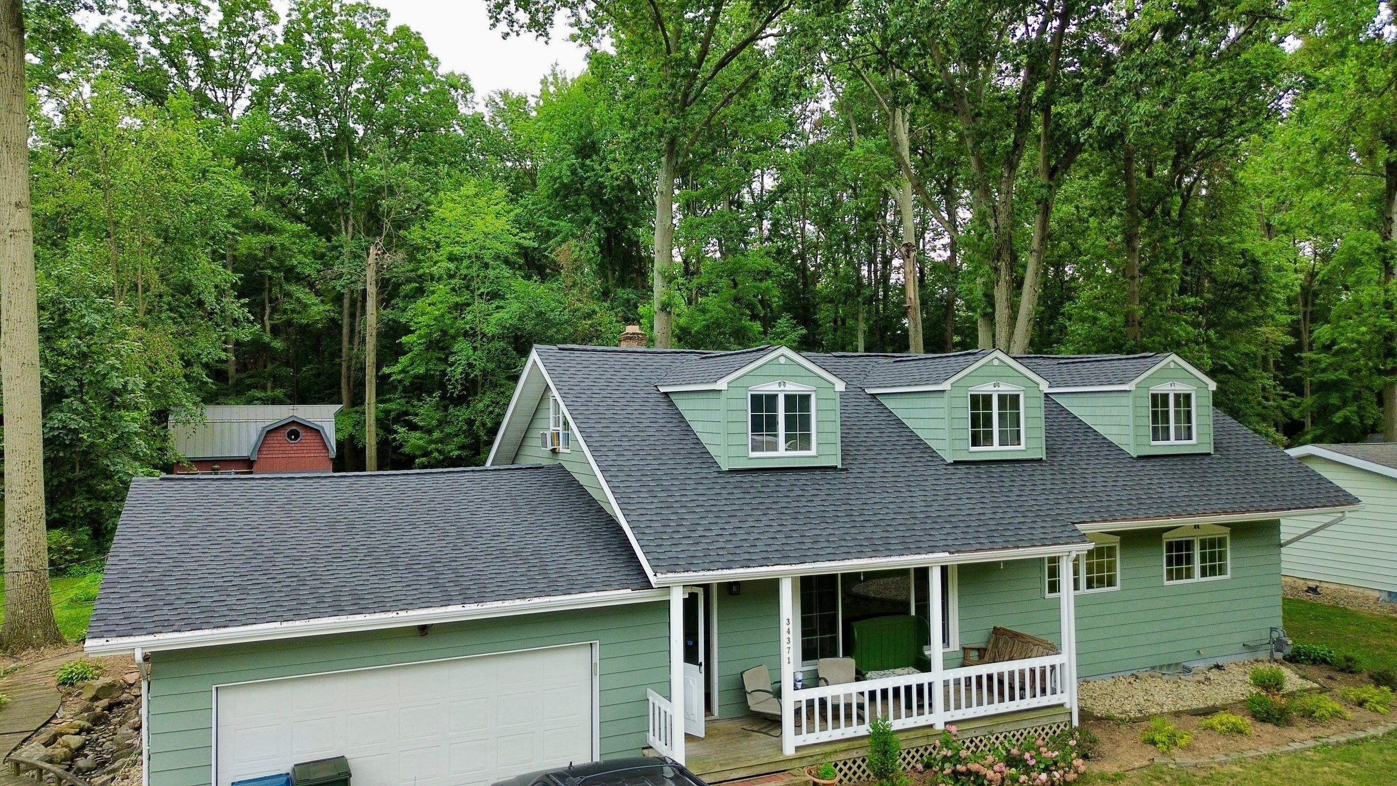 A green house with a porch, situated among tall trees and a nearby red shed.