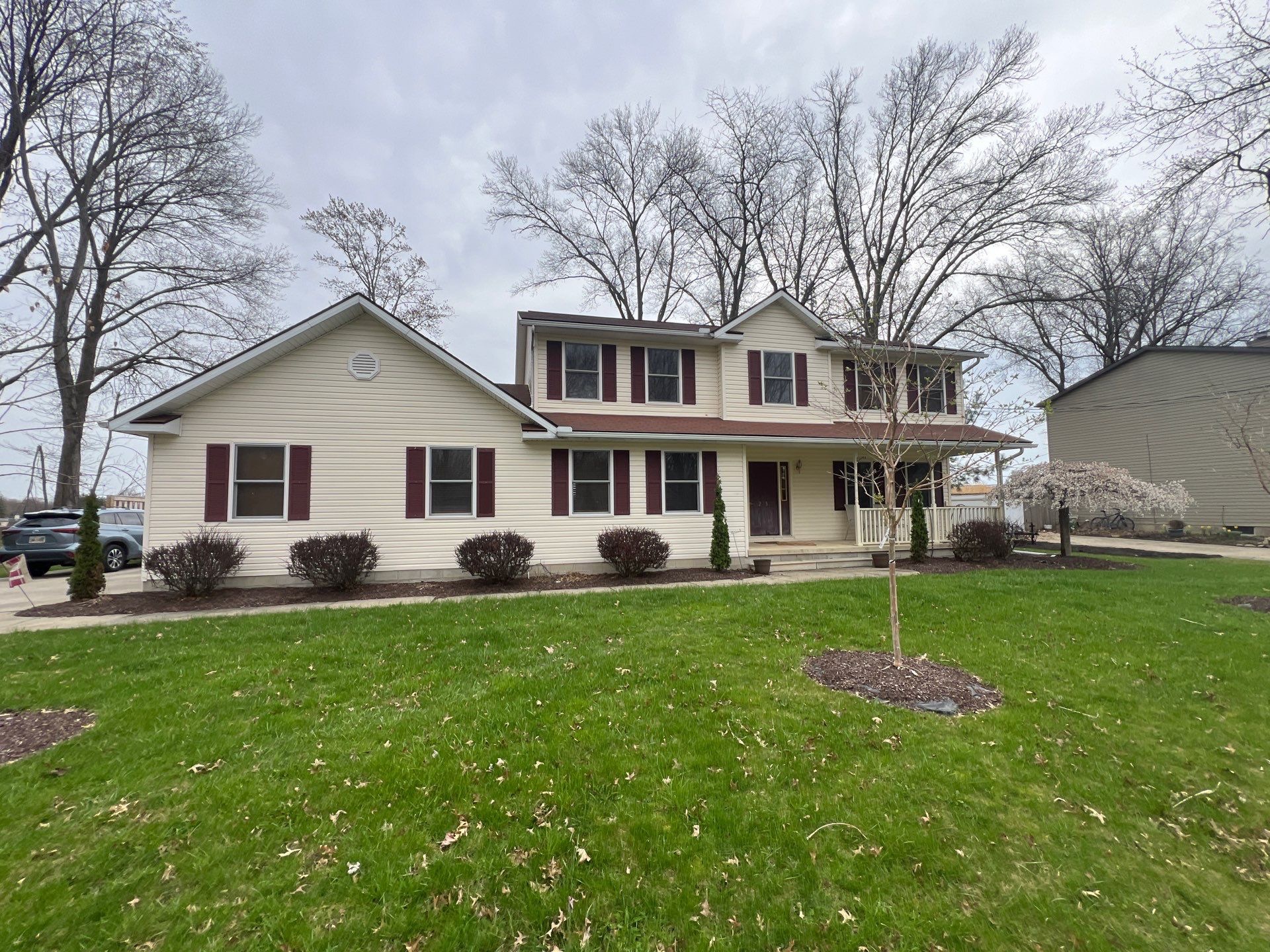 A two-story beige house with maroon shutters sits in a manicured lawn under a cloudy sky.
