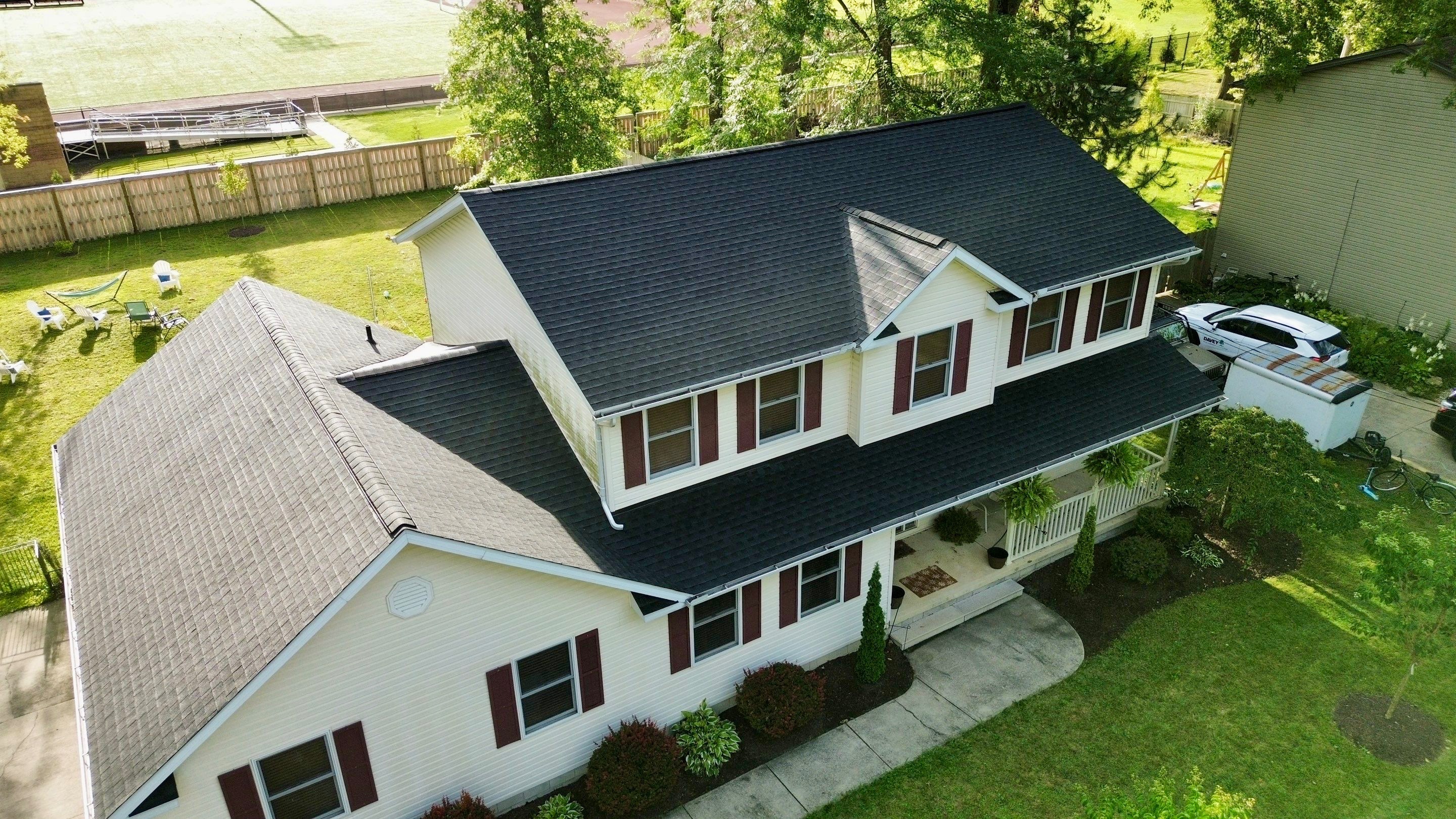 A two-story house with a black roof, white siding, and a green lawn, surrounded by trees and outdoor furniture.