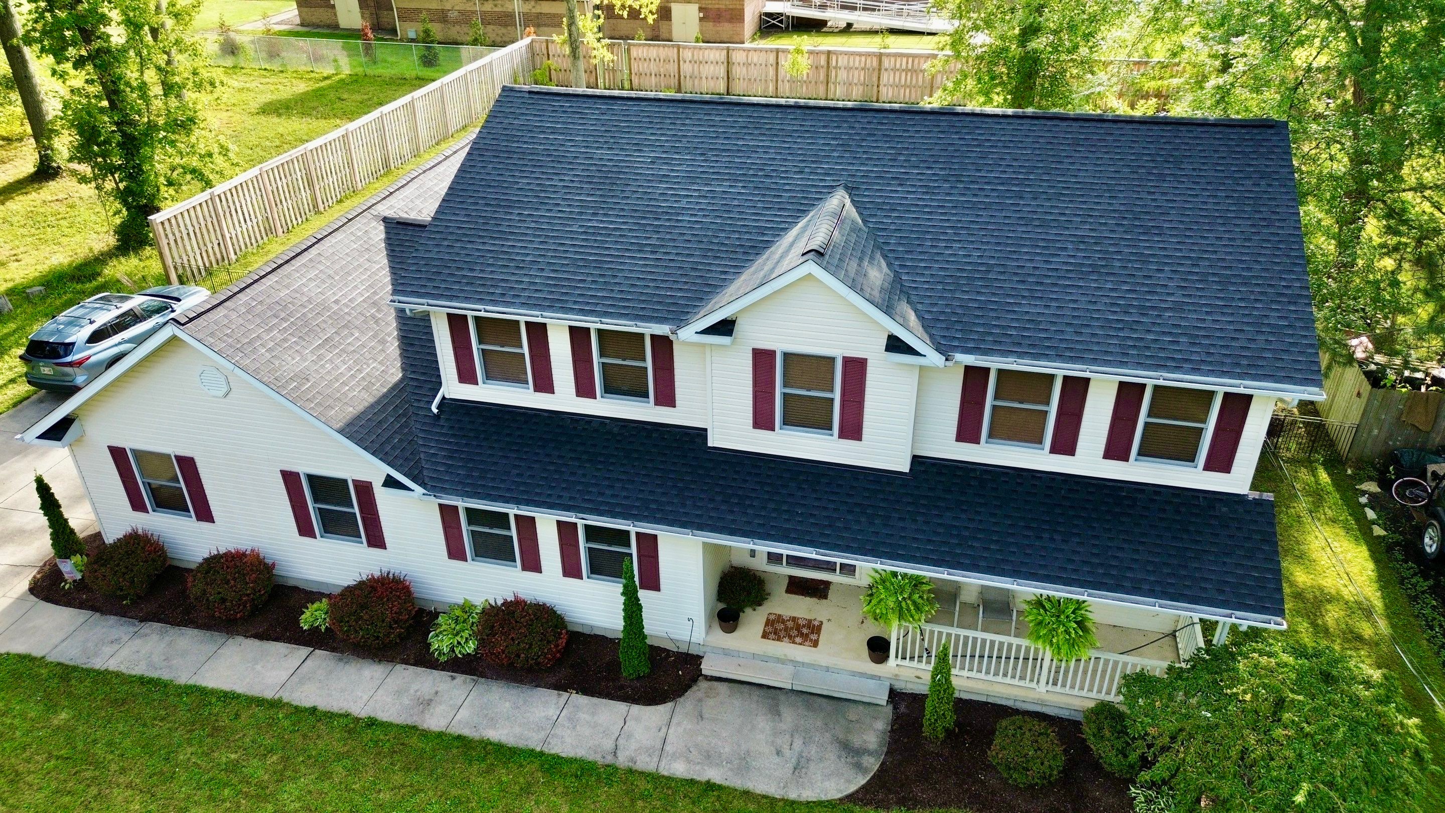 A two-story white house with a black roof and red shutters, surrounded by green grass and trees.