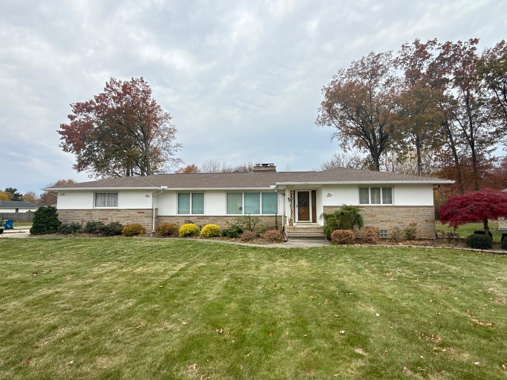 A single-story house with a stone foundation, surrounded by a grassy lawn and autumn trees.