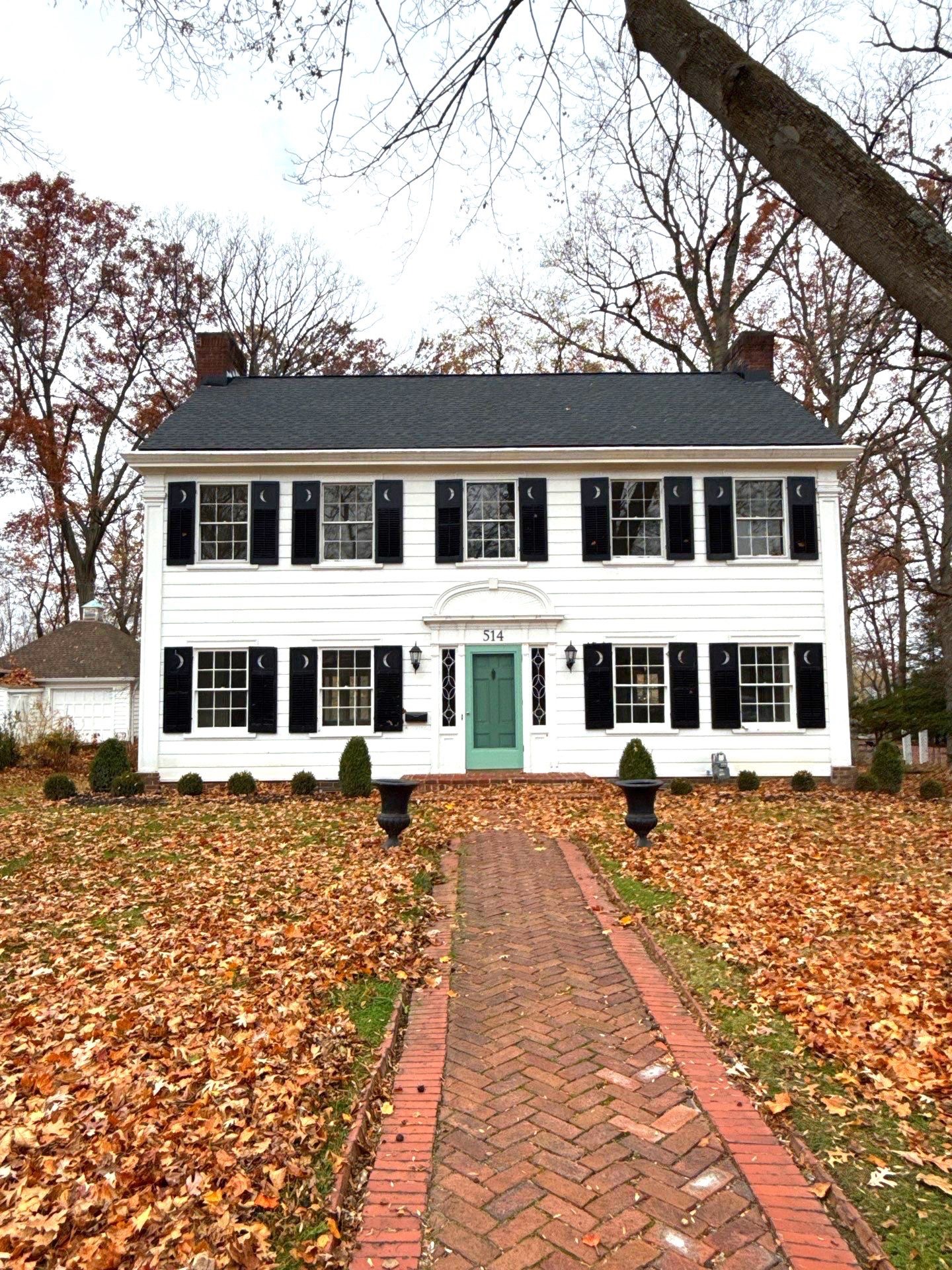 A two-story white house with black shutters and a green door, surrounded by fallen leaves and shrubs.