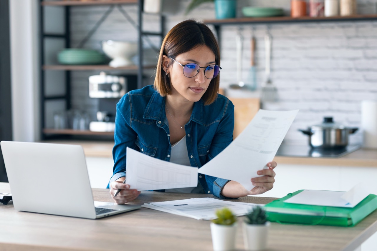 Young business woman working with computer while consulting some invoices and documents in the kitchen at home.