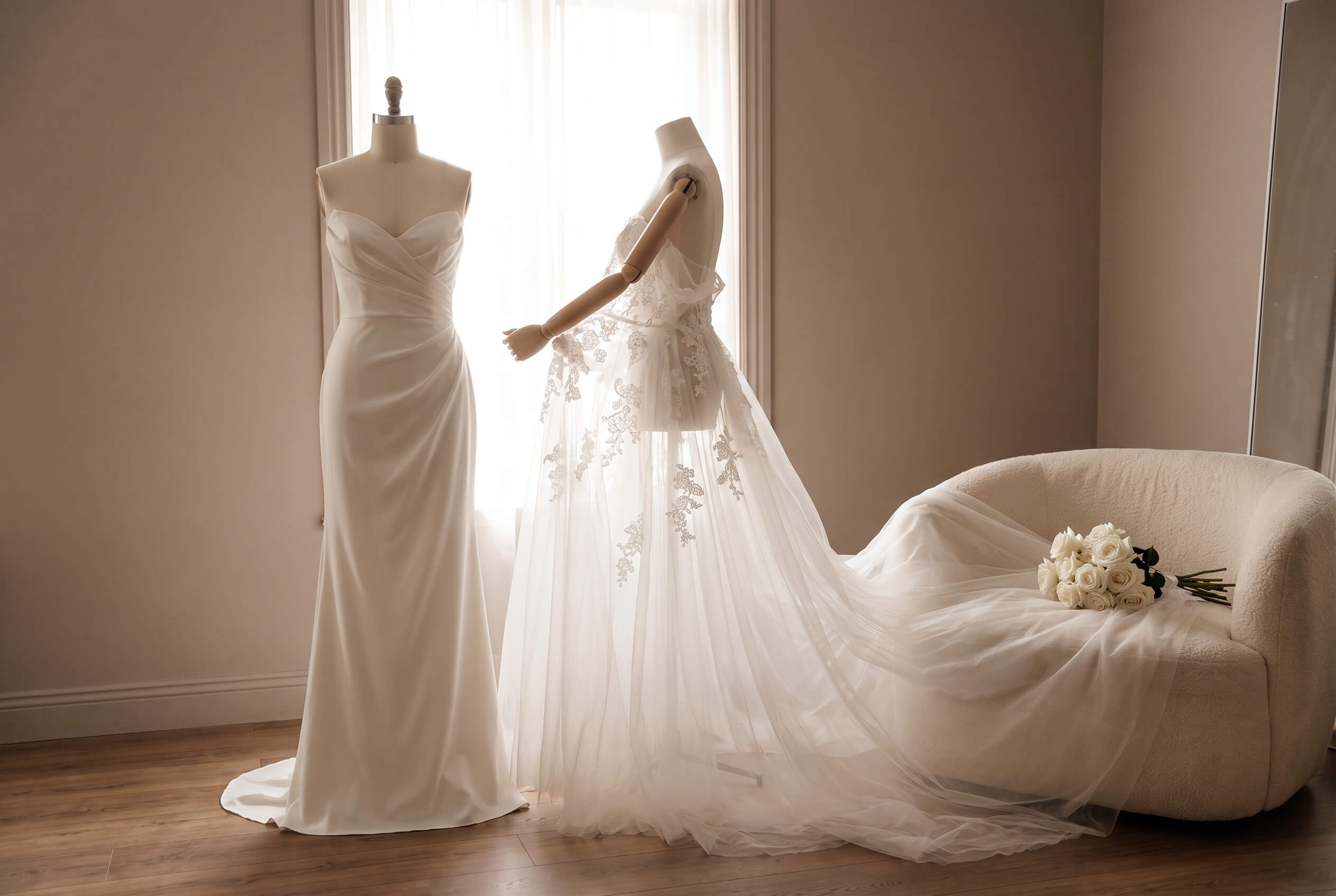 Two wedding dresses displayed on mannequins in a softly lit room; one is a sleek satin gown and the other features lace details and a long tulle train extending onto a nearby cream-colored curved chair with a bouquet of white roses on it.