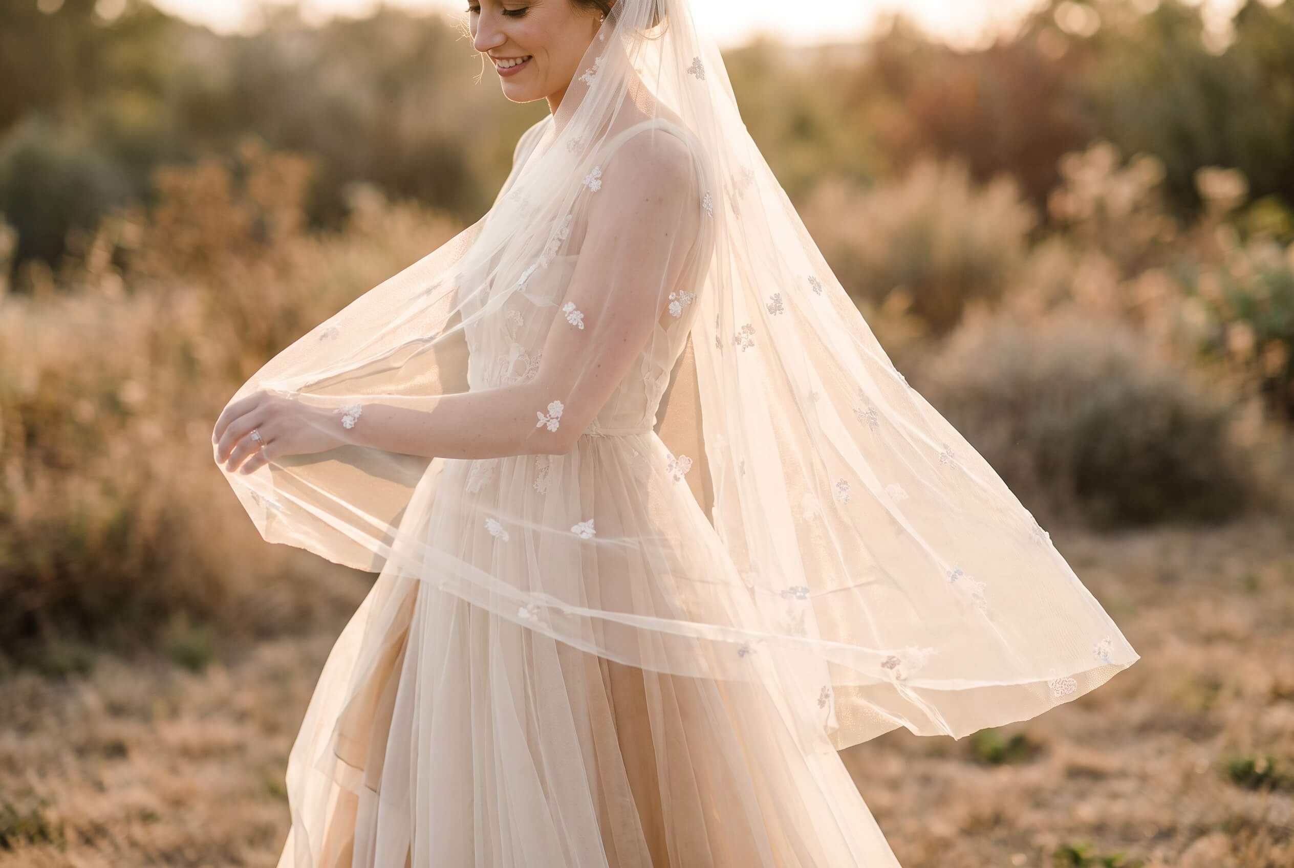 Side view of a smiling bride wearing a flowing wedding dress and a sheer veil with floral embroidery, standing outdoors in warm, golden light.