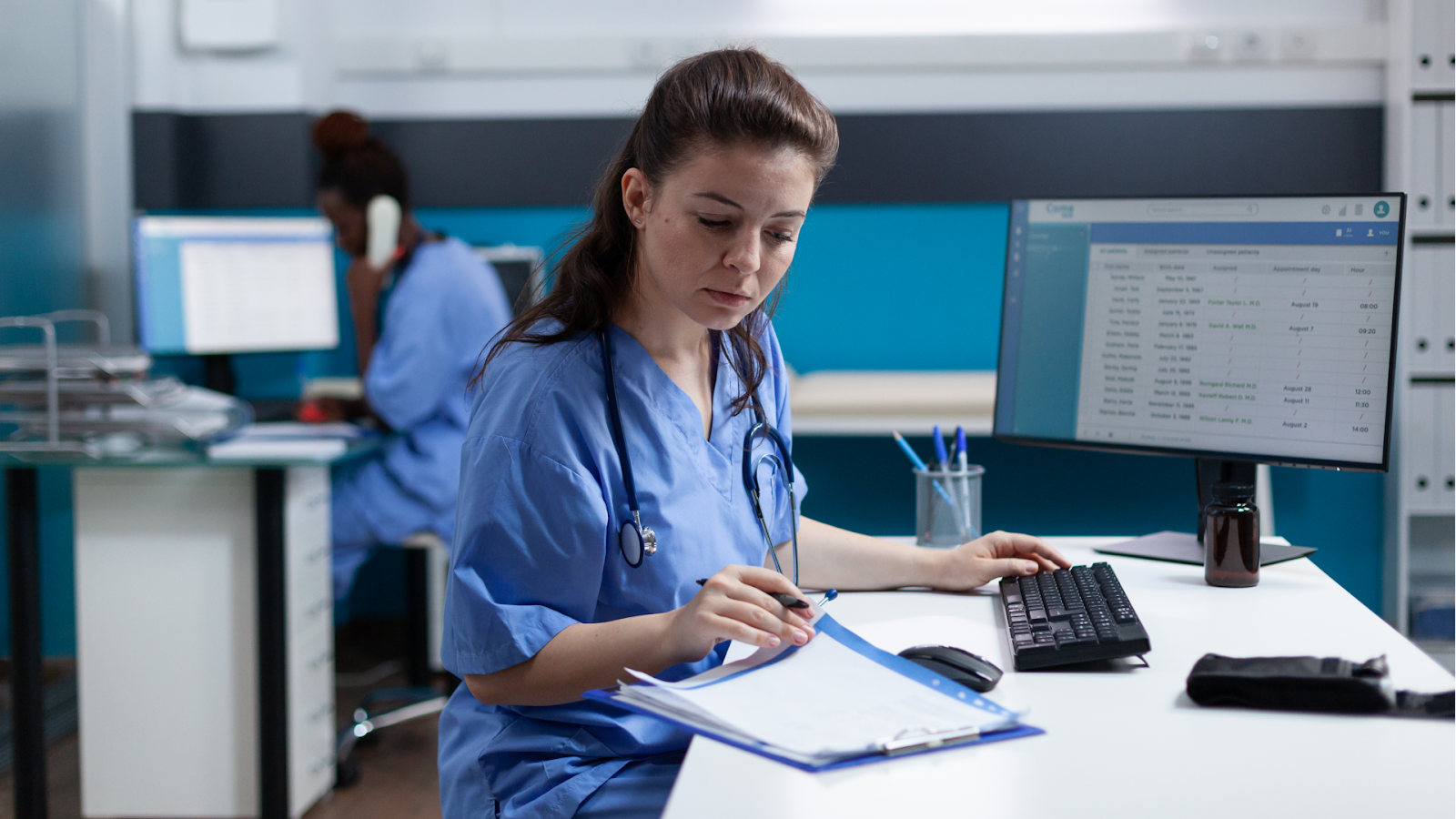 A nurse in blue scrubs reviews patient records on a clipboard while working at a computer in a hospital office.