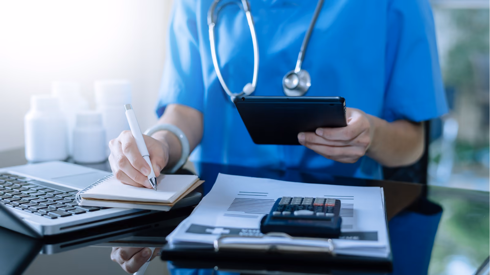 A nurse in blue scrubs writes notes in a notebook while holding a tablet with a laptop and a calculator on the desk.