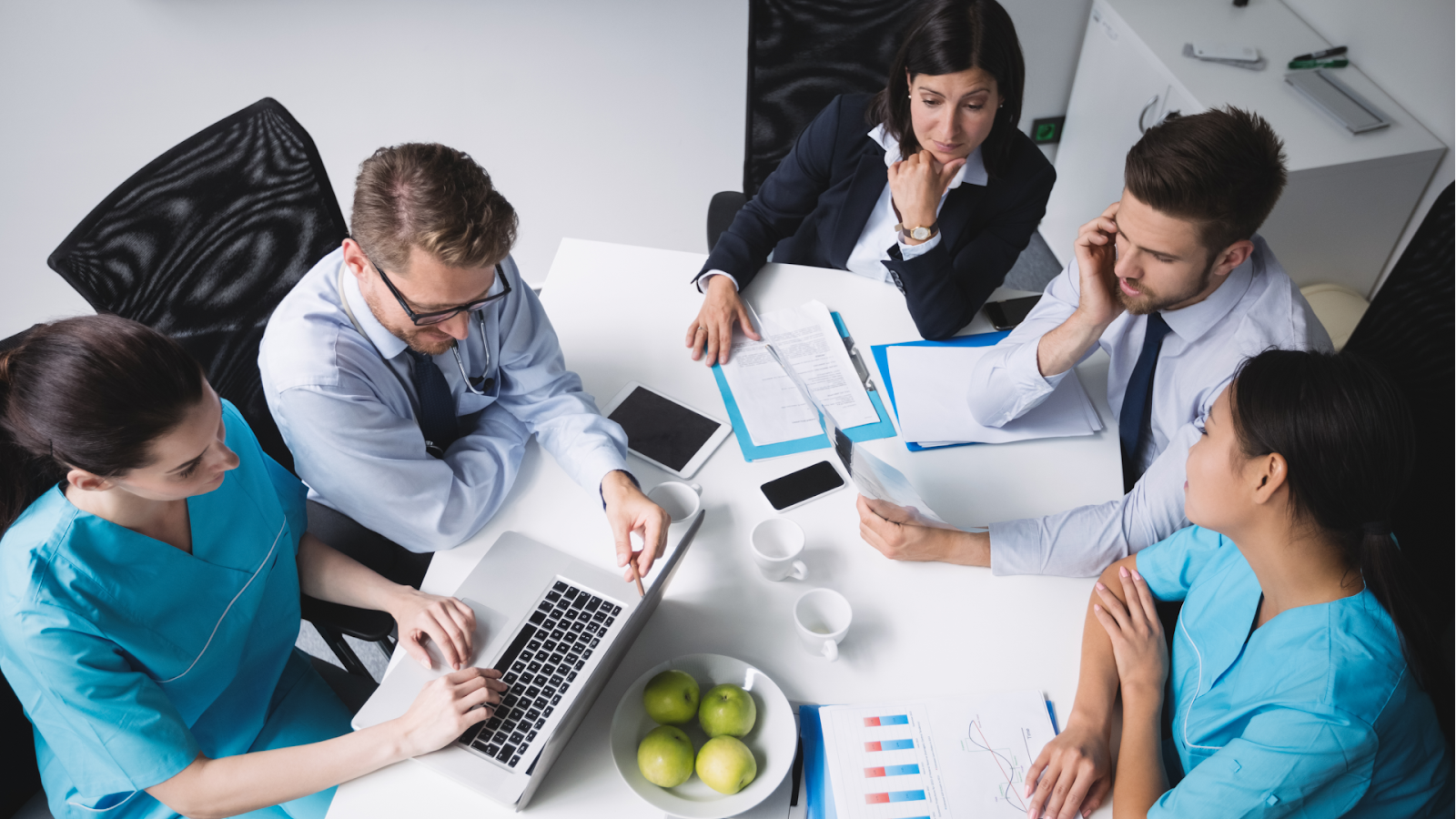 A team of healthcare professionals and administrators sits around a table reviewing medical charts and financial graphs.