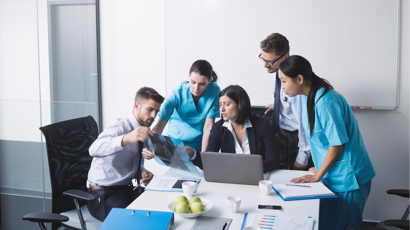 A group of doctors and nurses gather around a conference table, examining an X-ray while discussing patient care, with a laptop and medical files in front of them.