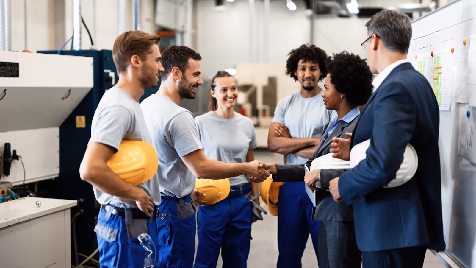 A group of workers in safety gear and uniform standing around two business professionals in a manufacturing facility.
