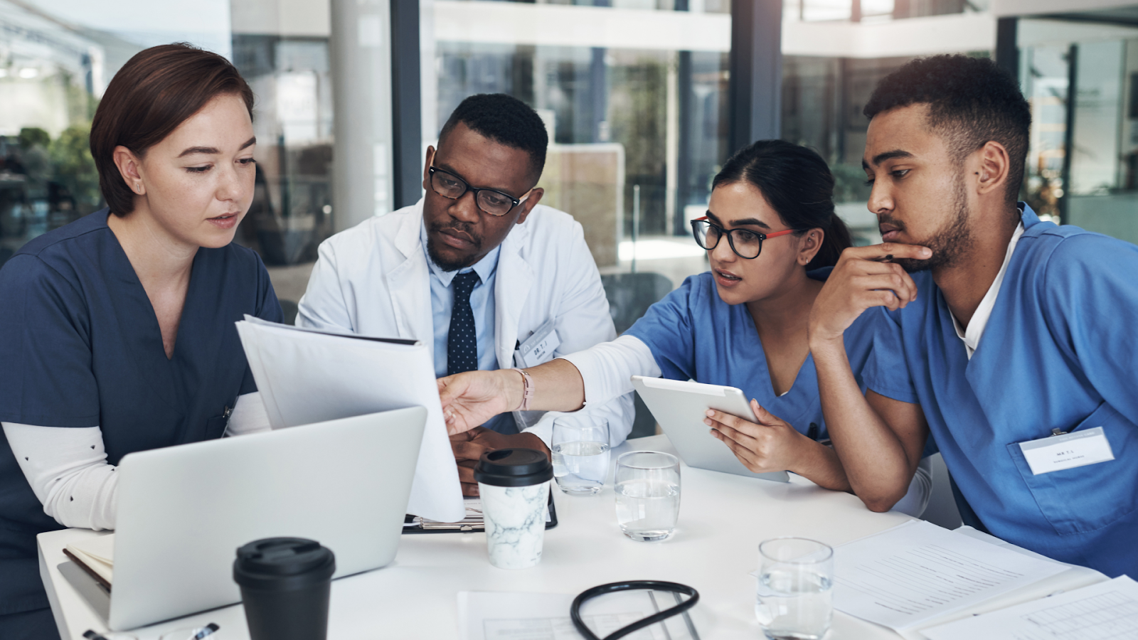 Four medical professionals gathered around a table with a laptop and papers, discussing documents and a digital tablet.