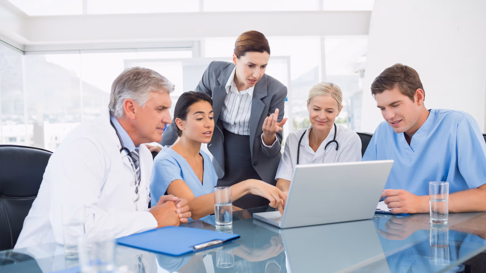 Five healthcare professionals are seated around a table with a laptop, pointing at the screen and discussing a project