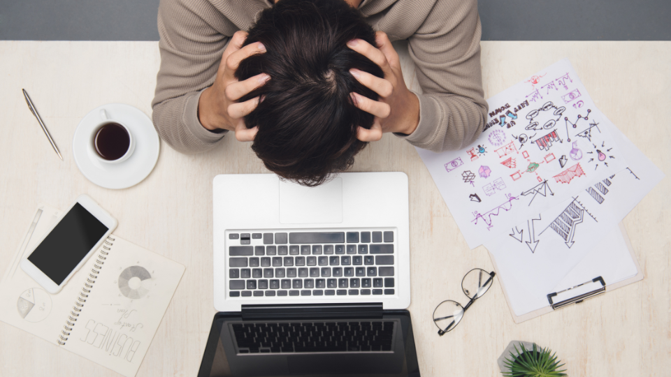 An overhead image of a stressed employee holding their head over a laptop surrounded by scattered papers.