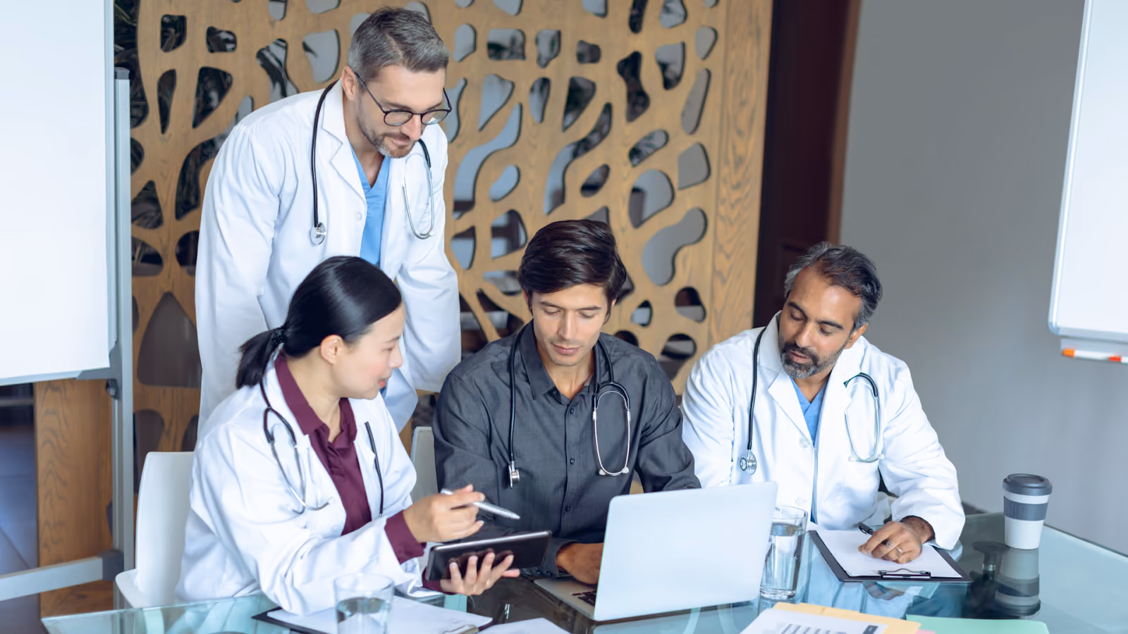 A group of four medical professionals gathered around a desk, focused on a laptop and a tablet for a collaborative consultation.