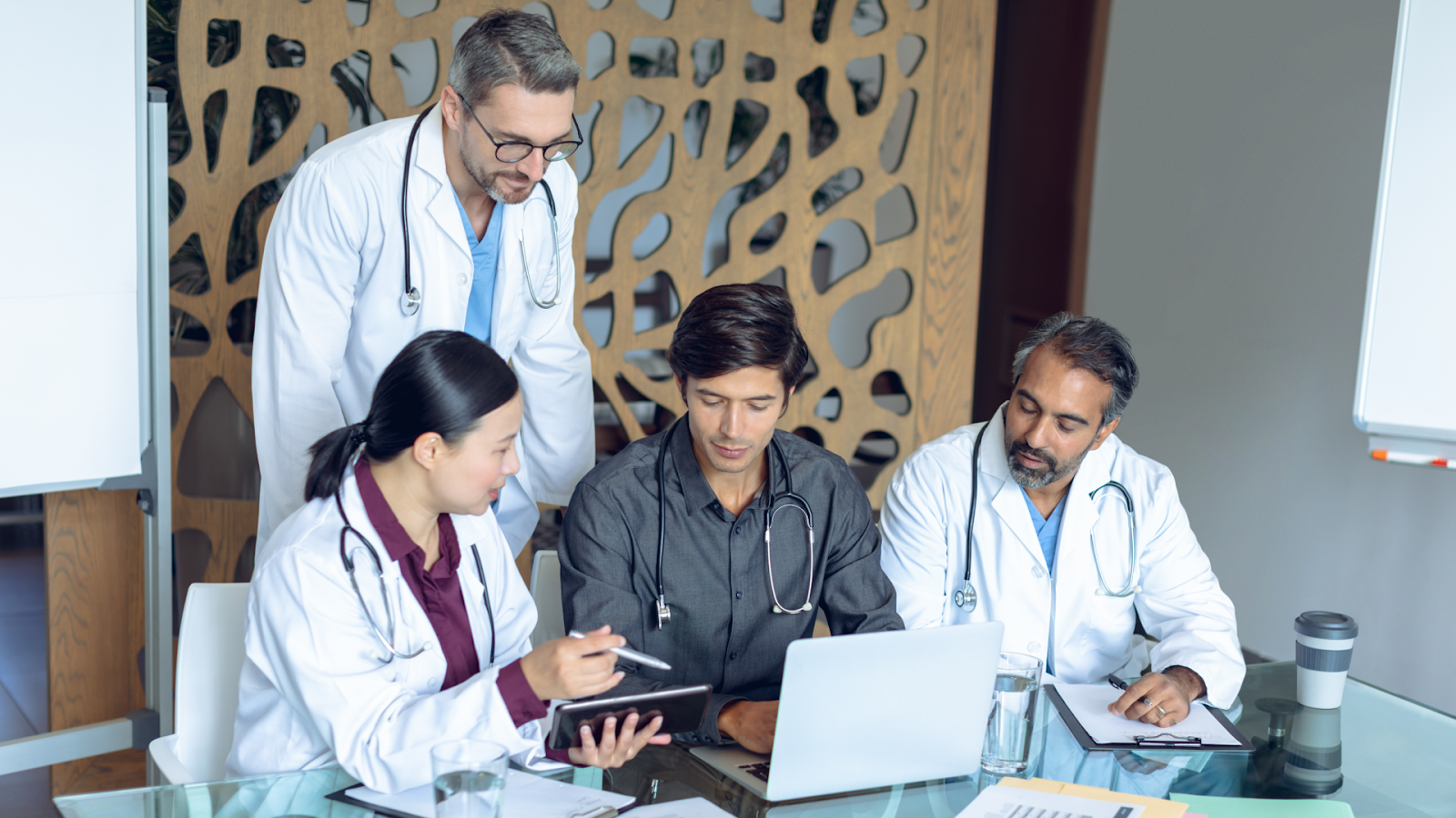 A group of four medical professionals gathered around a desk, focused on a laptop and a tablet for a collaborative consultation.