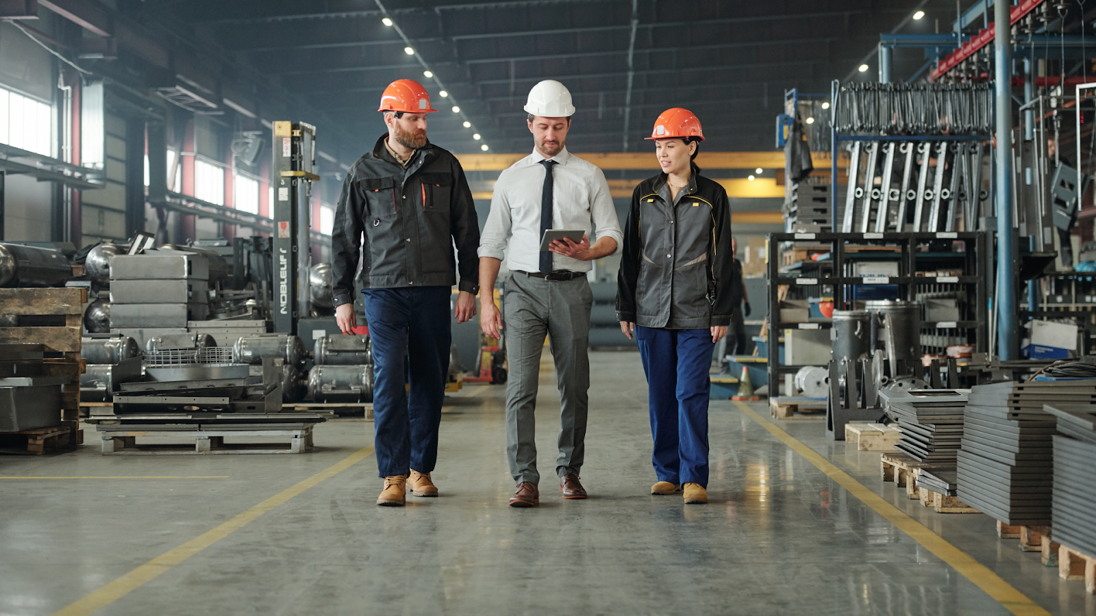 Three people walking and talking in the aisle of a large industrial warehouse or factory.